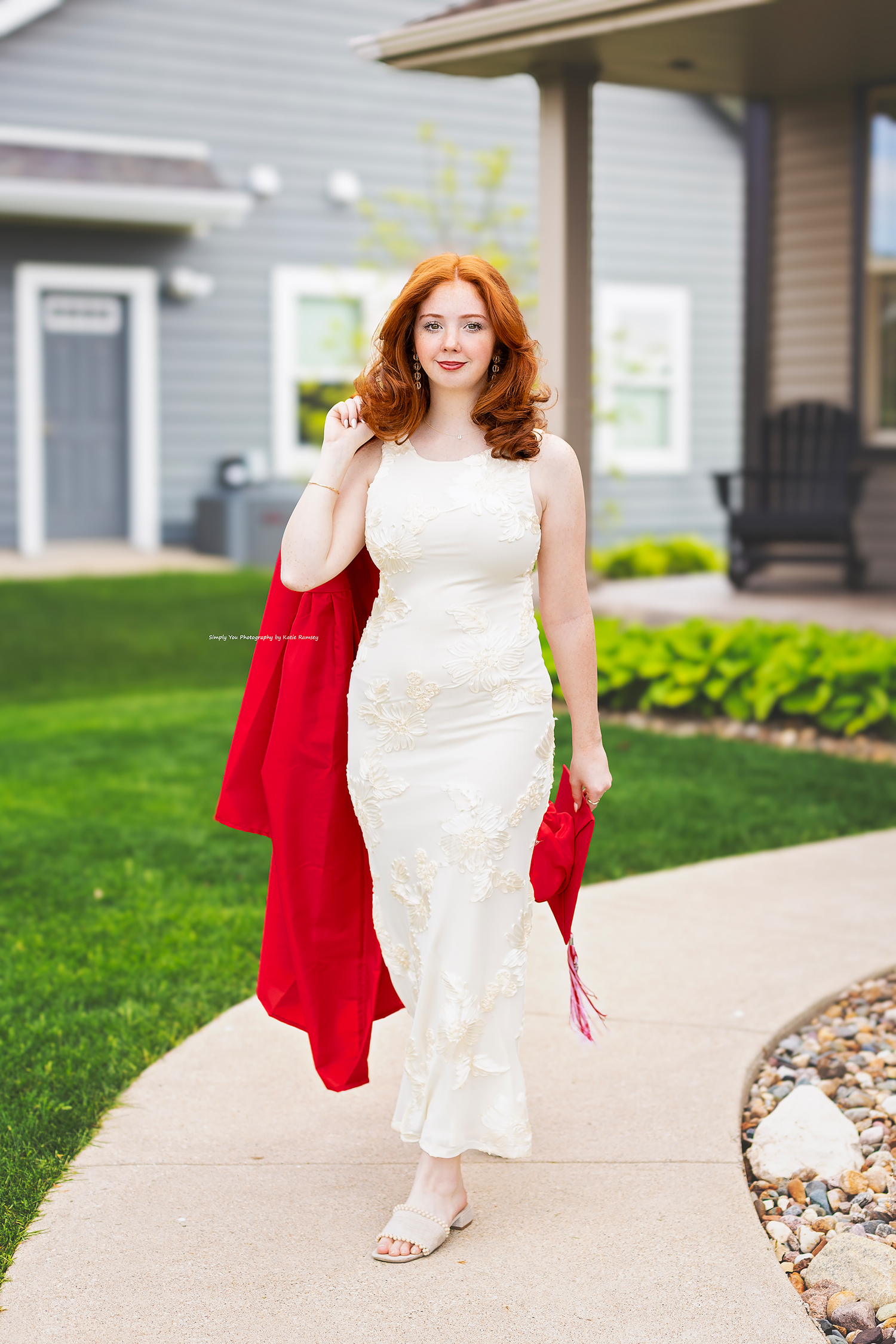 Young woman with red hair in white lace dress carrying red graduation gown on a sidewalk.