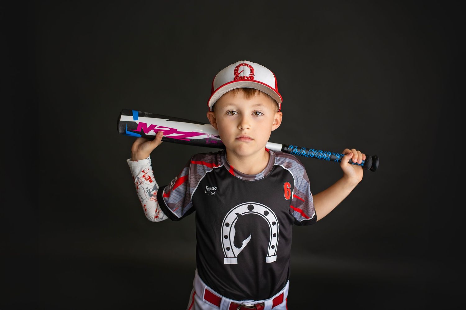Boy in baseball uniform holds bat across shoulders with serious expression in studio photo.