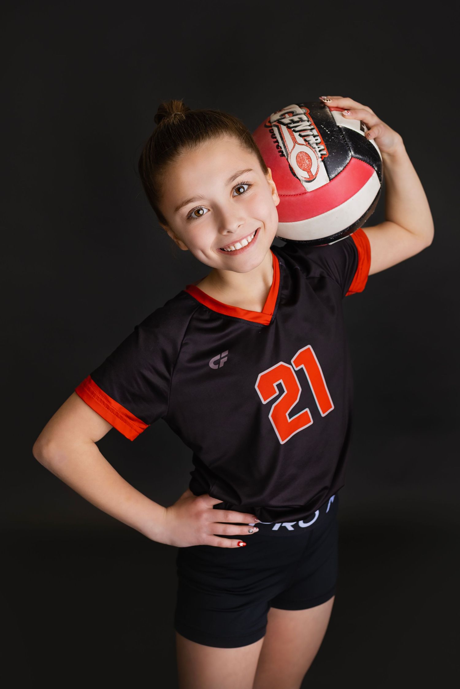 Smiling girl in number 21 black and red volleyball jersey holding ball on shoulder with hand on hip.