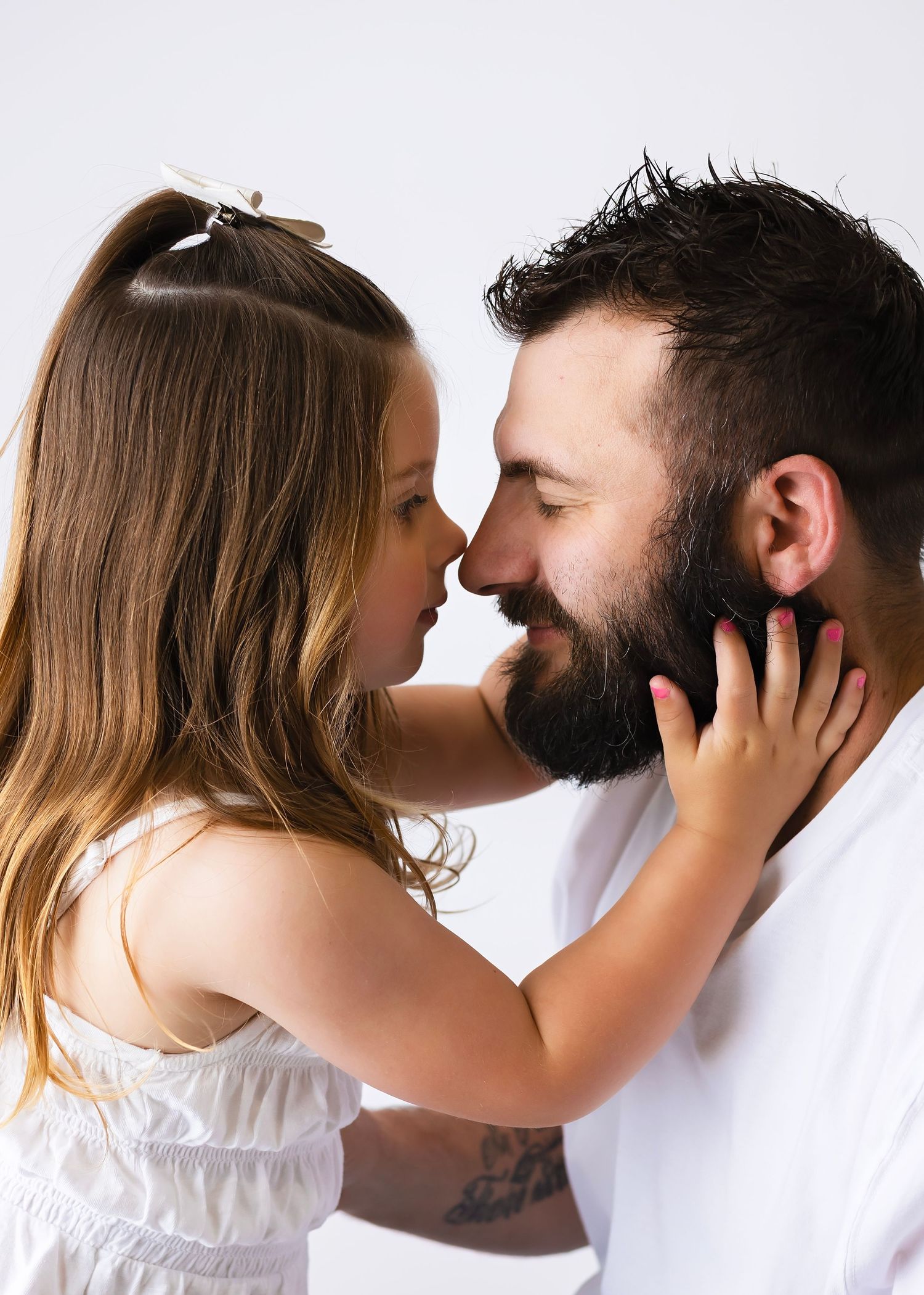 A bearded father and young daughter share a tender nose-to-nose moment, both smiling in white outfits.