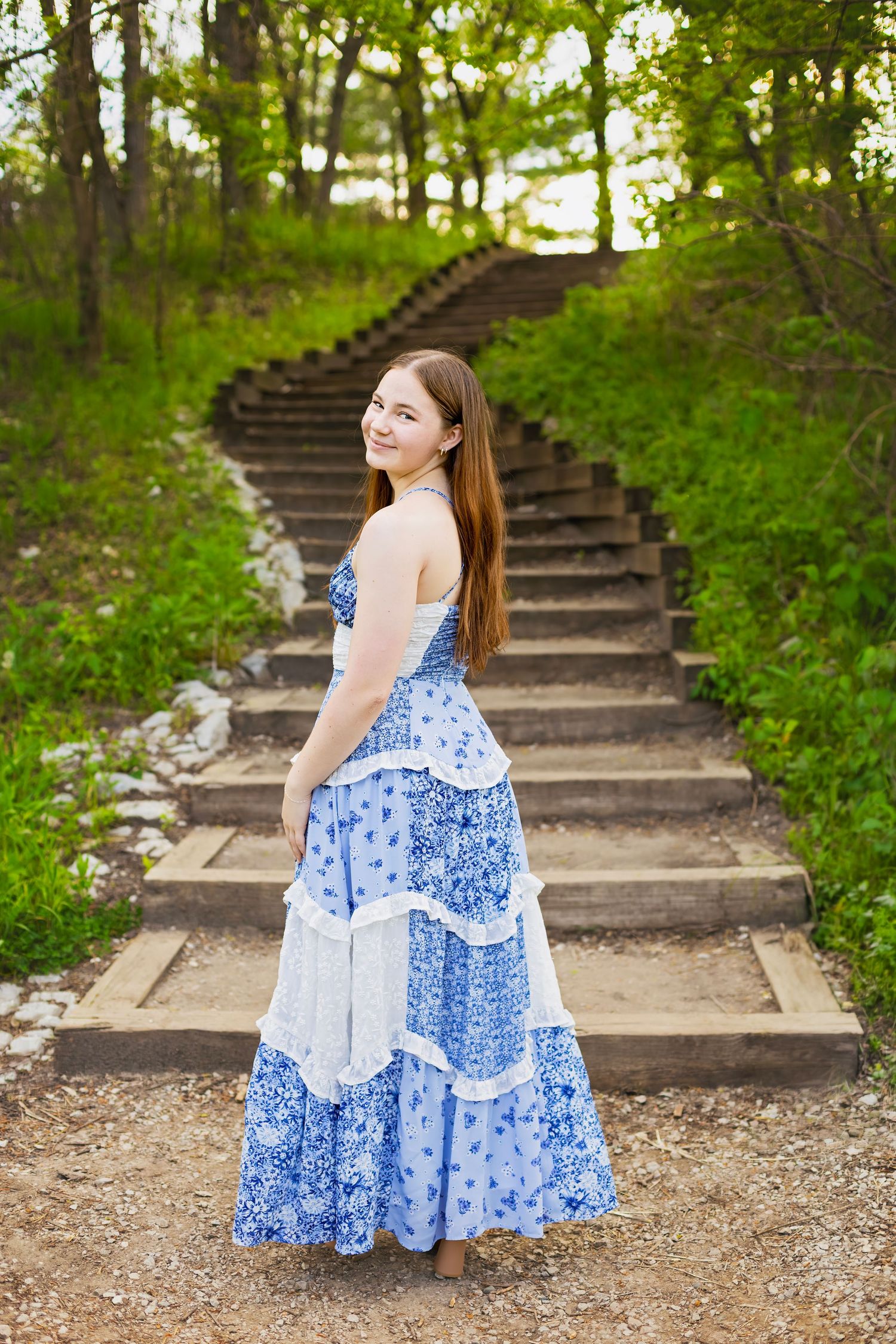 Young woman in blue floral maxi dress standing on stone steps in a green forest.