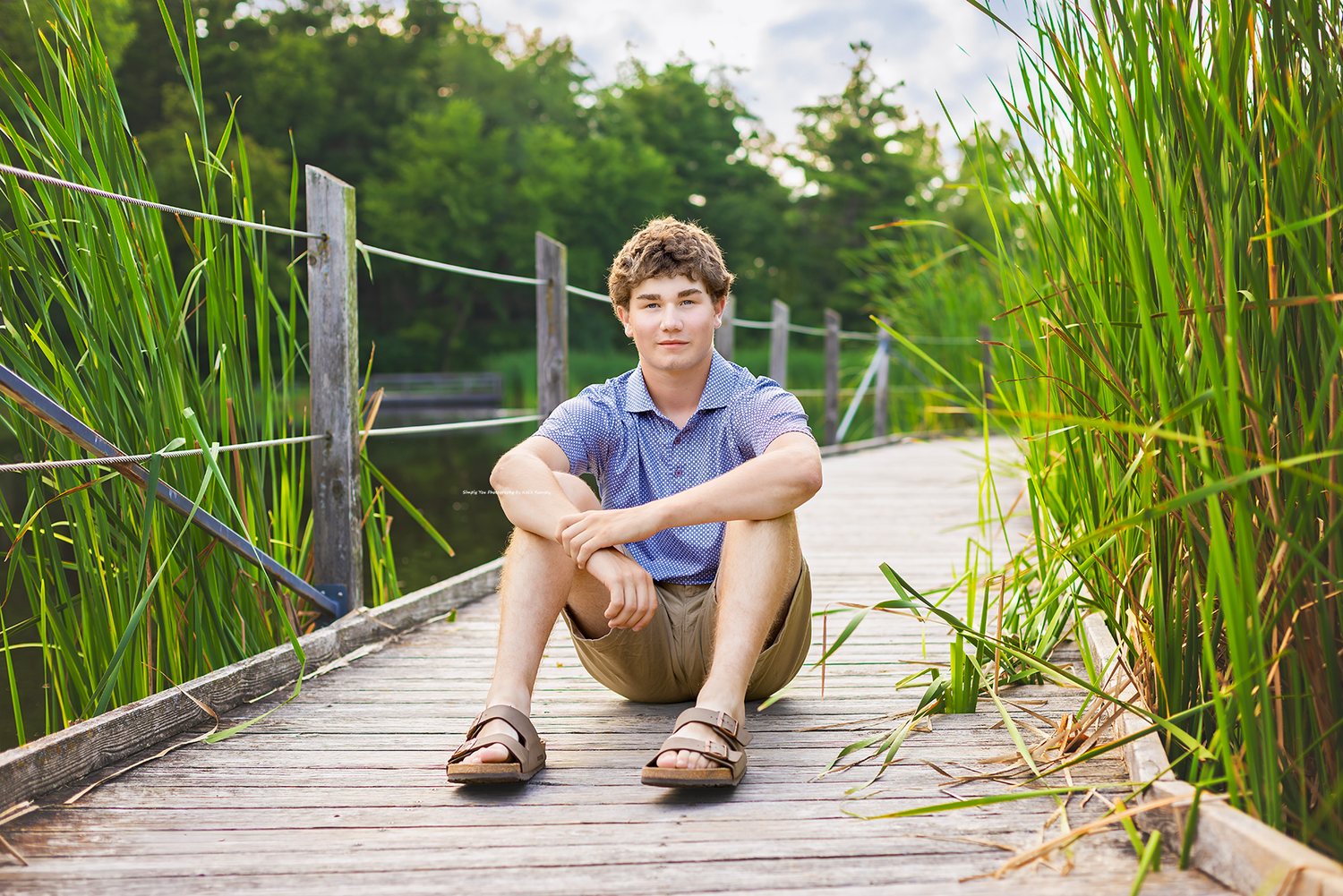 Teen boy sitting cross-legged on a wooden dock surrounded by tall green reeds near a calm lake on a sunny day.