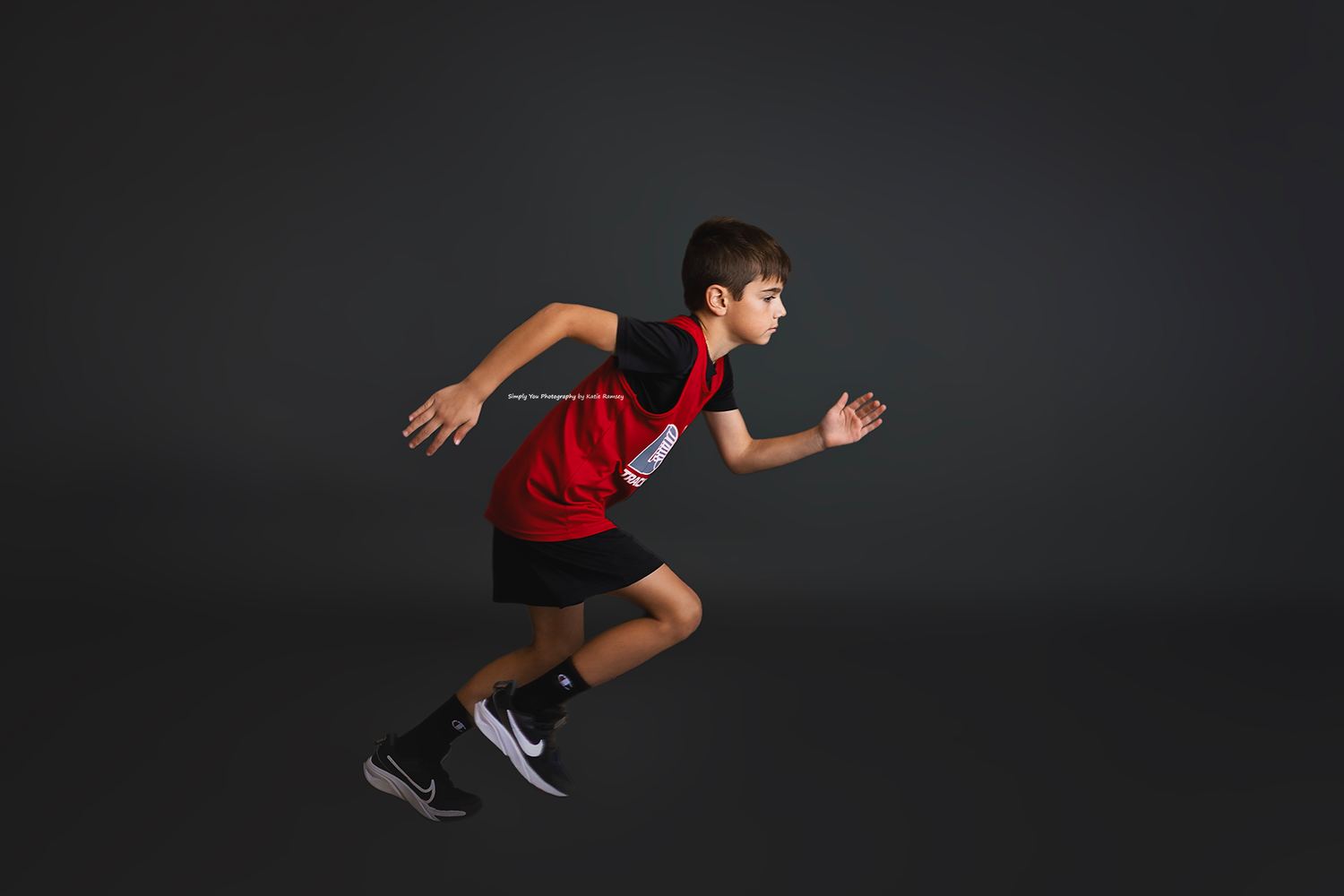 Boy in red and black athletic wear sprinting dynamically against a dark gray studio background.
