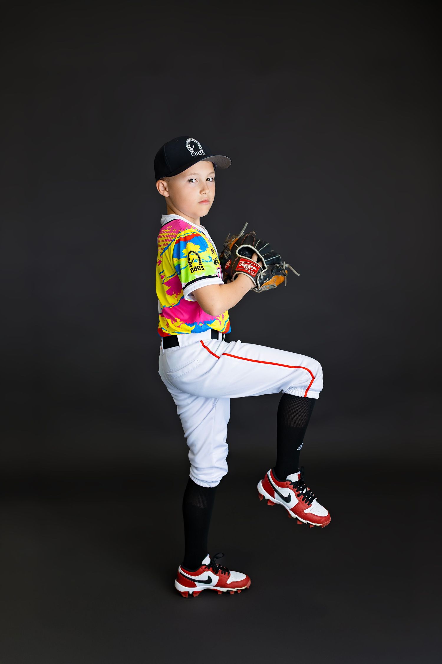Young baseball player in tie-dye jersey assumes pitching stance with glove raised wearing white pants and red cleats.