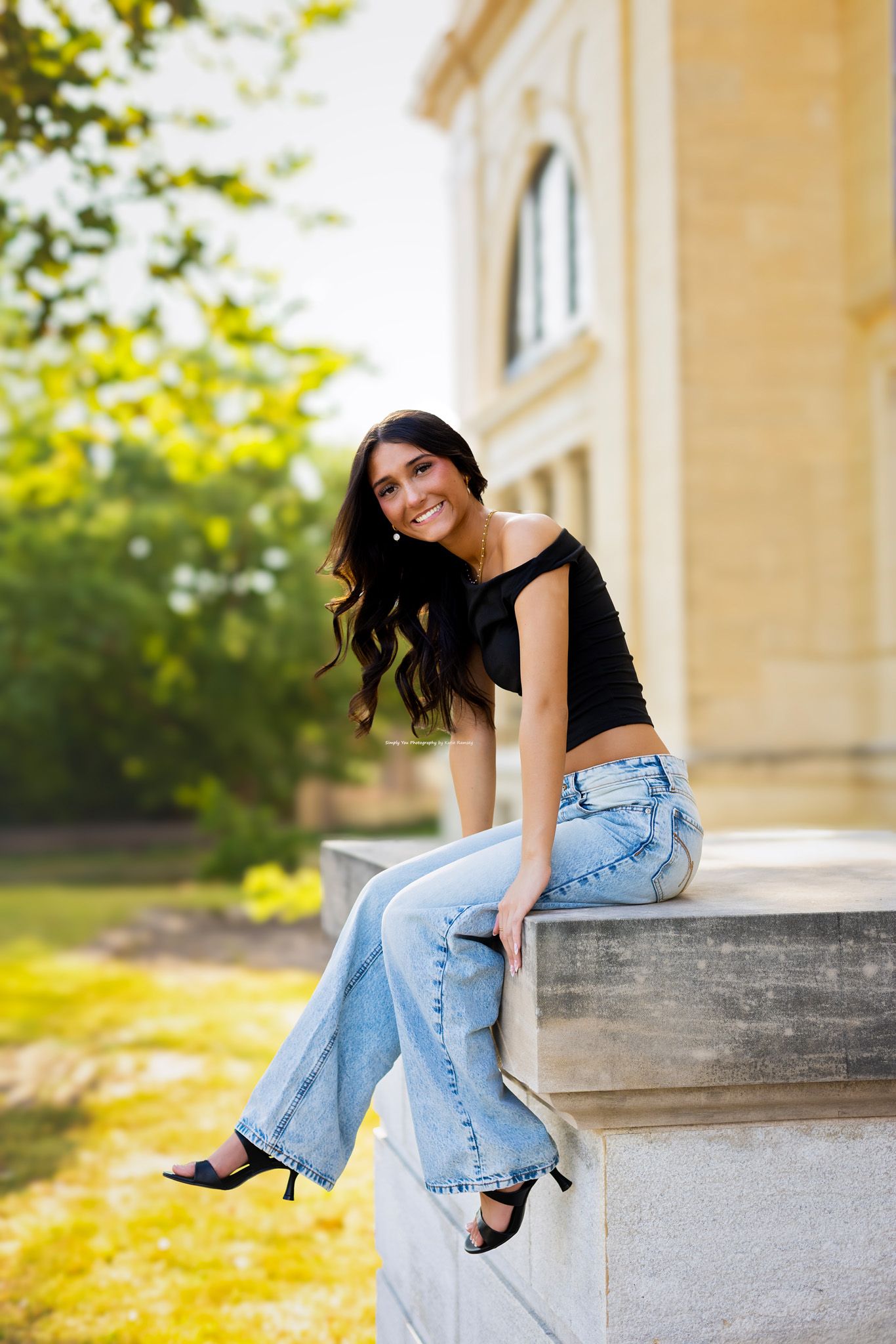 Young woman in black crop top and wide-leg jeans sitting on stone ledge outdoors, smiling confidently.
