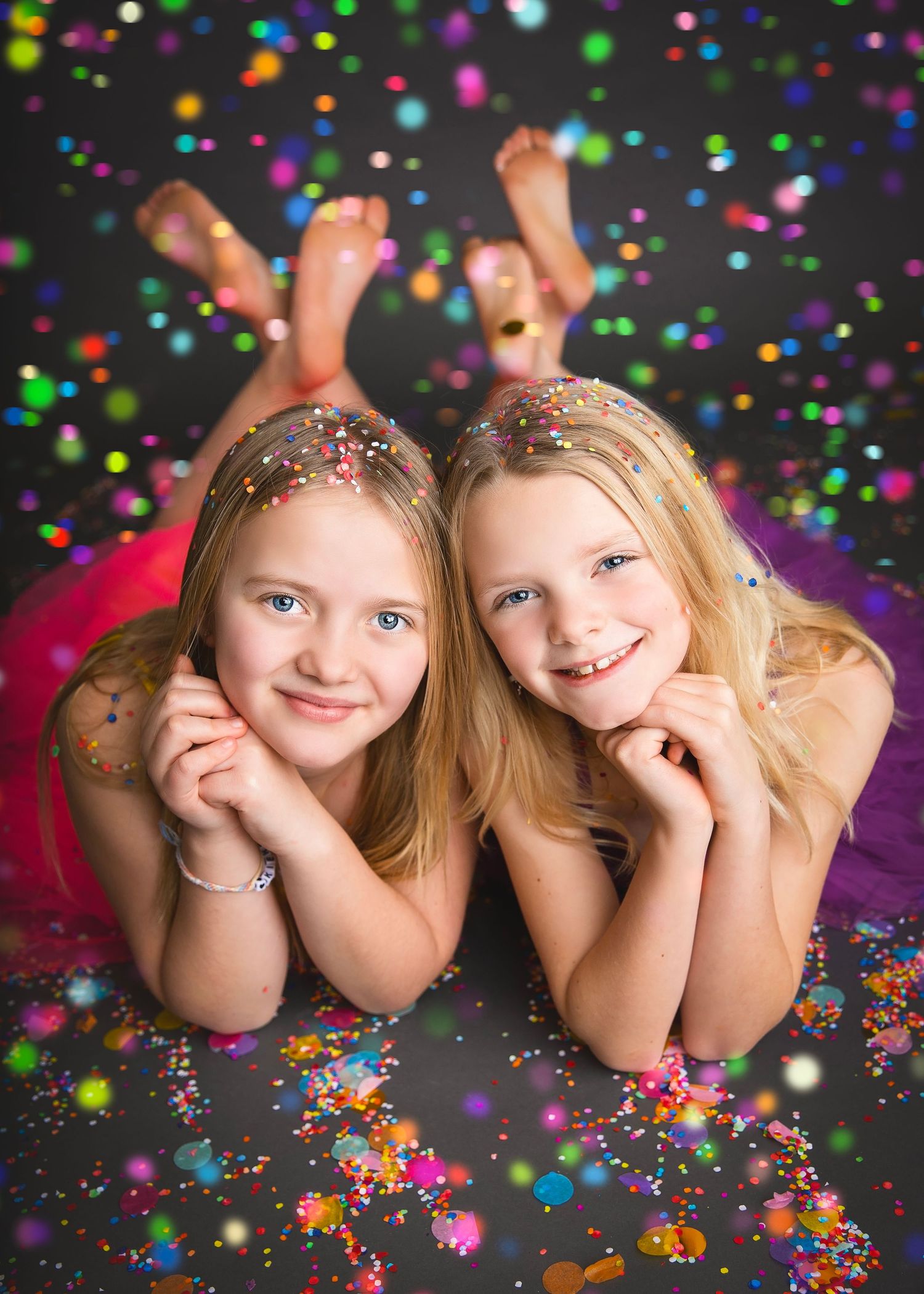 Two blonde sisters lying down smiling with colorful glitter bokeh lights surrounding them.