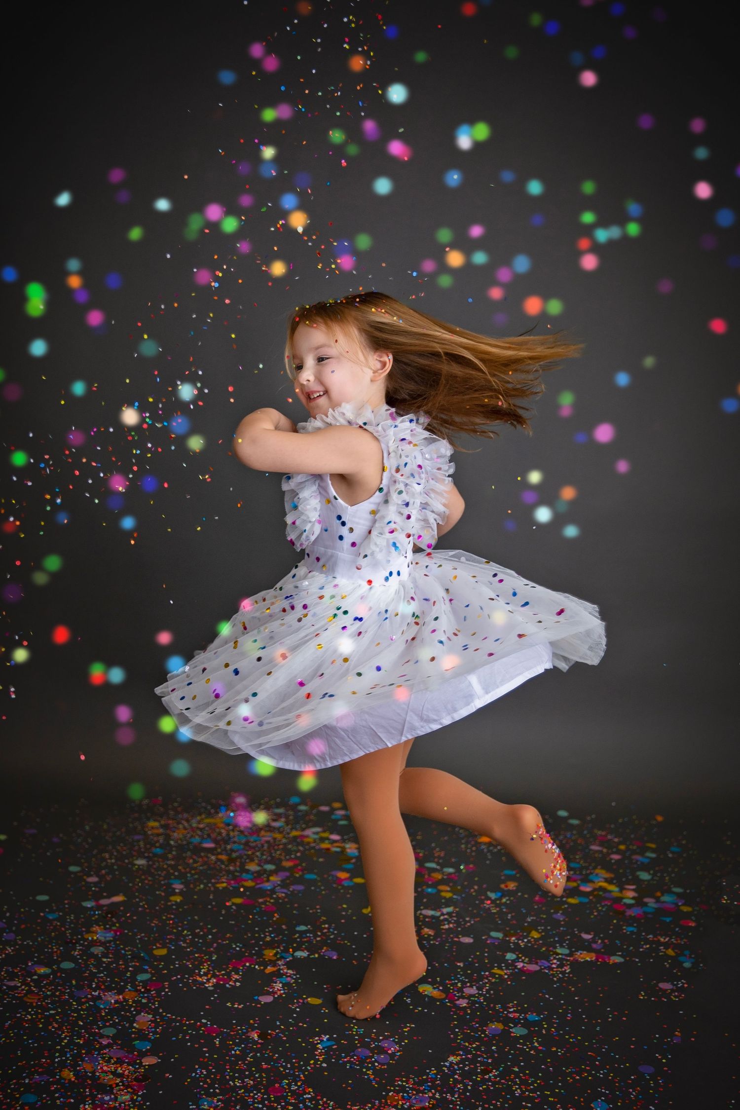 A joyful young girl in a white dress spins barefoot amid falling colorful confetti in a studio photo.
