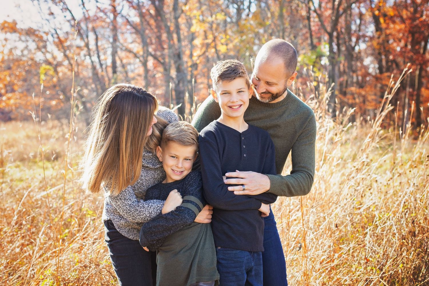 Family of four embracing in autumn field with golden foliage.
