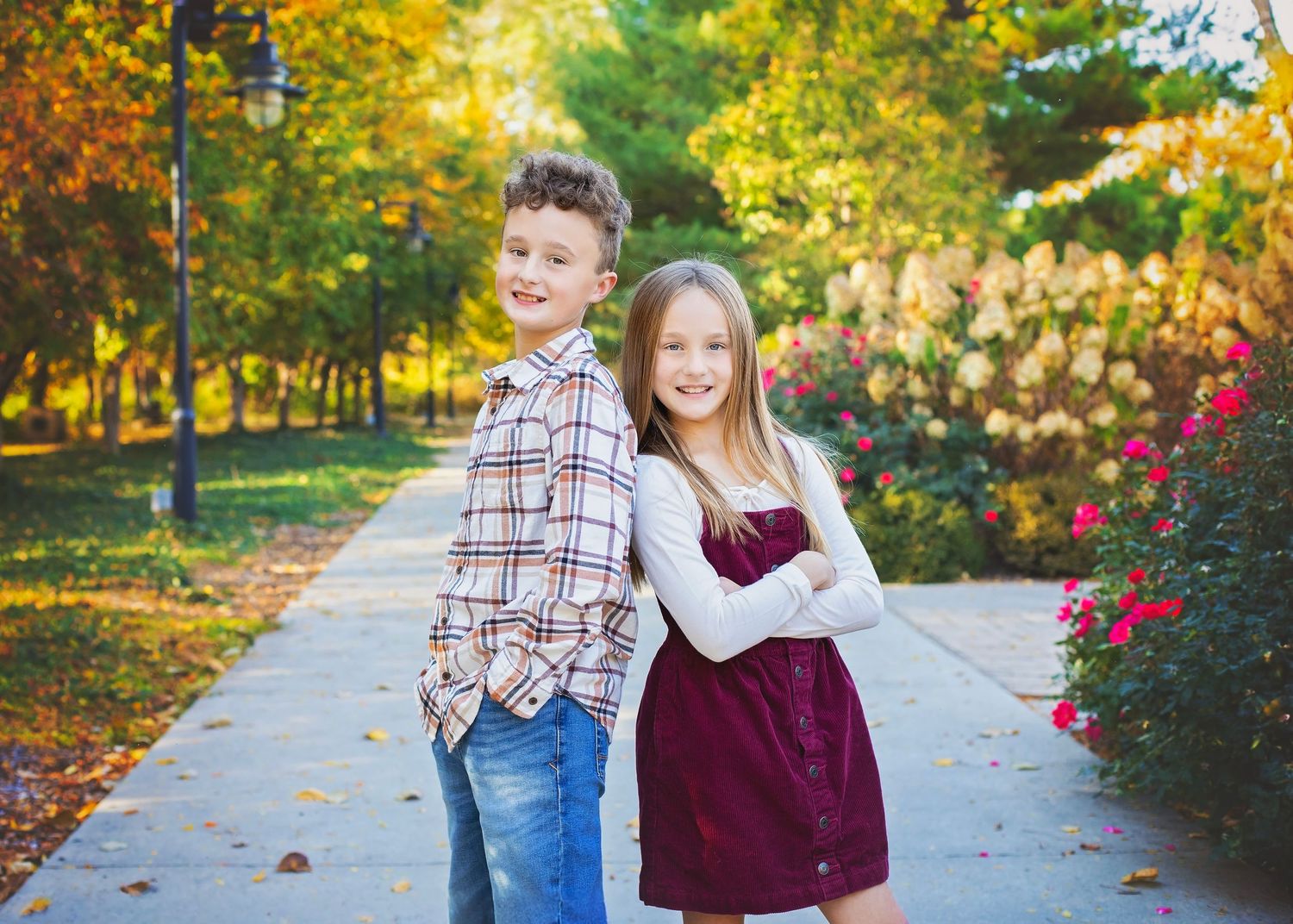 Two children standing back-to-back on a park path with fall foliage behind.