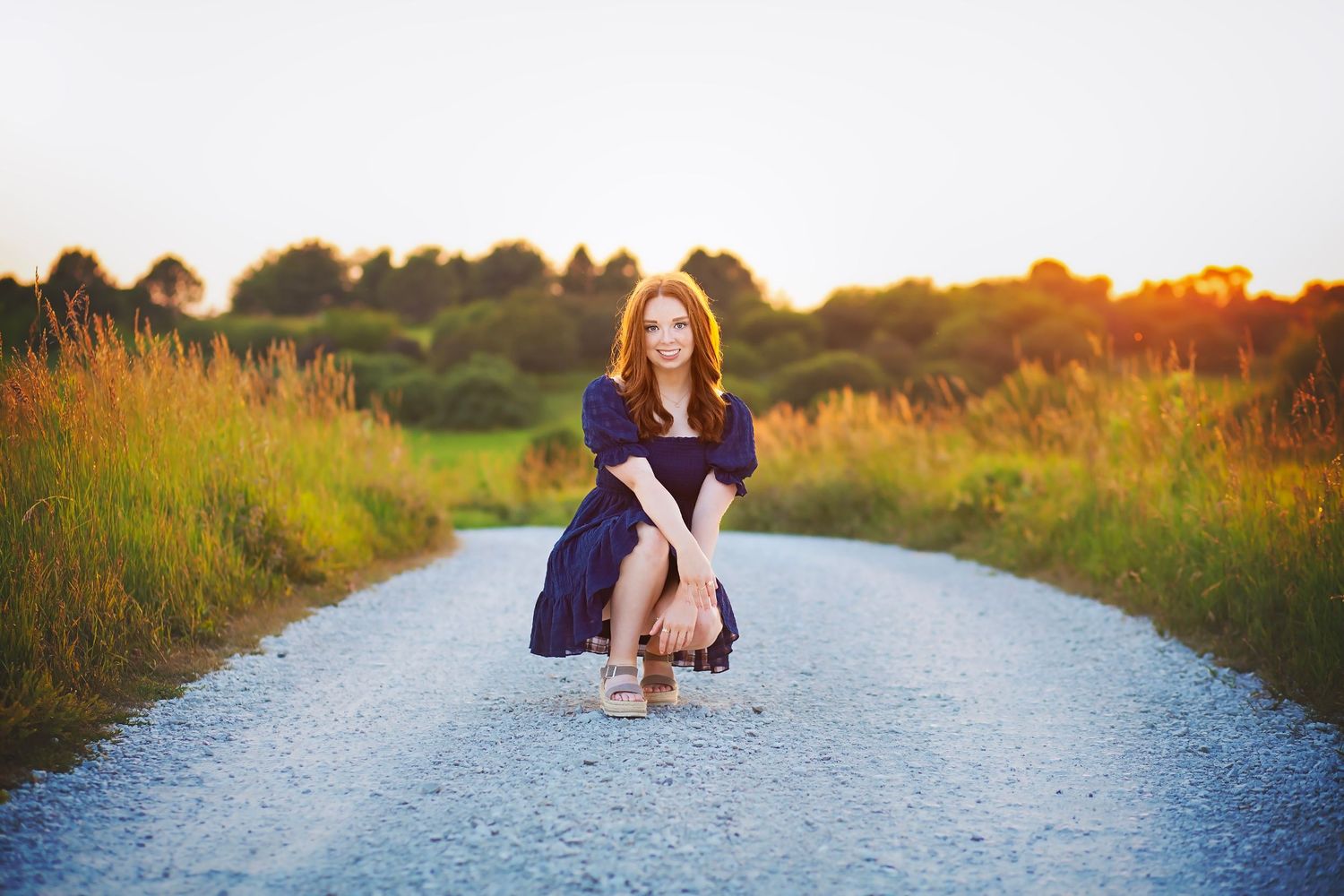 Redhead teen girl in navy dress crouches on rural gravel road surrounded by golden sunset meadow light.