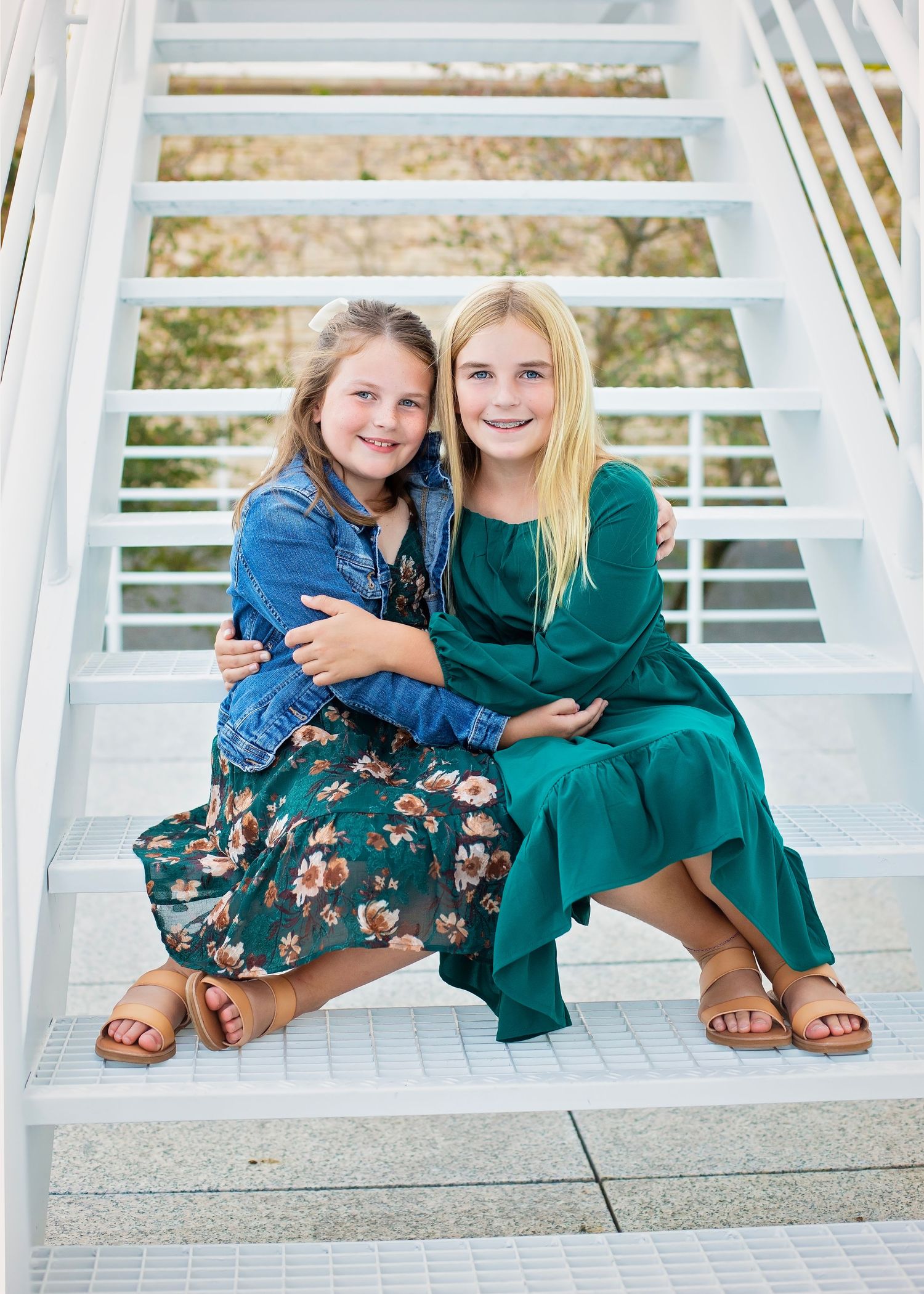 Two sisters sitting on white outdoor stairs, hugging and smiling, wearing green and floral dresses with sandals.