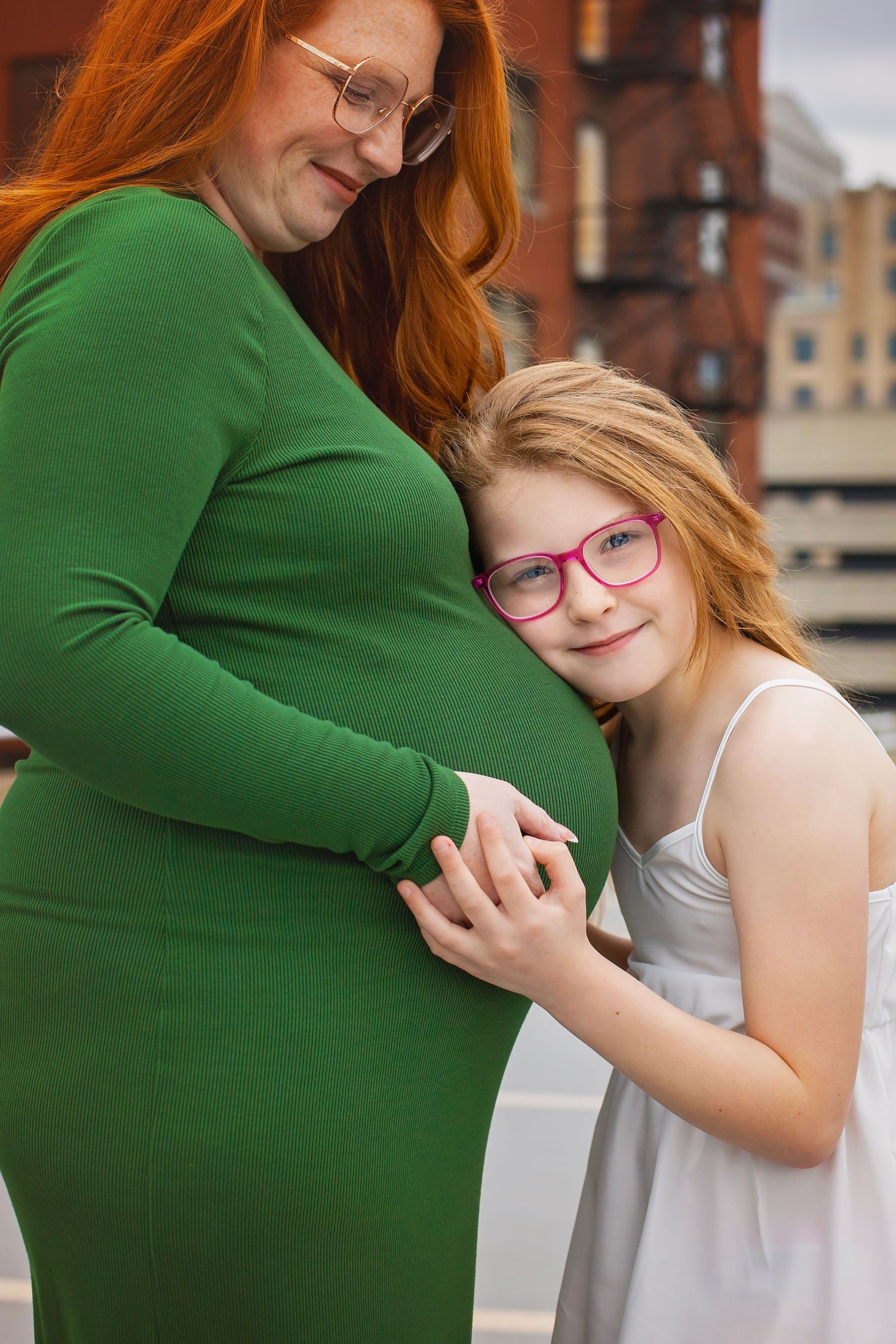Young red-haired girl with pink glasses hugging pregnant mothers green dress baby bump outdoors.
