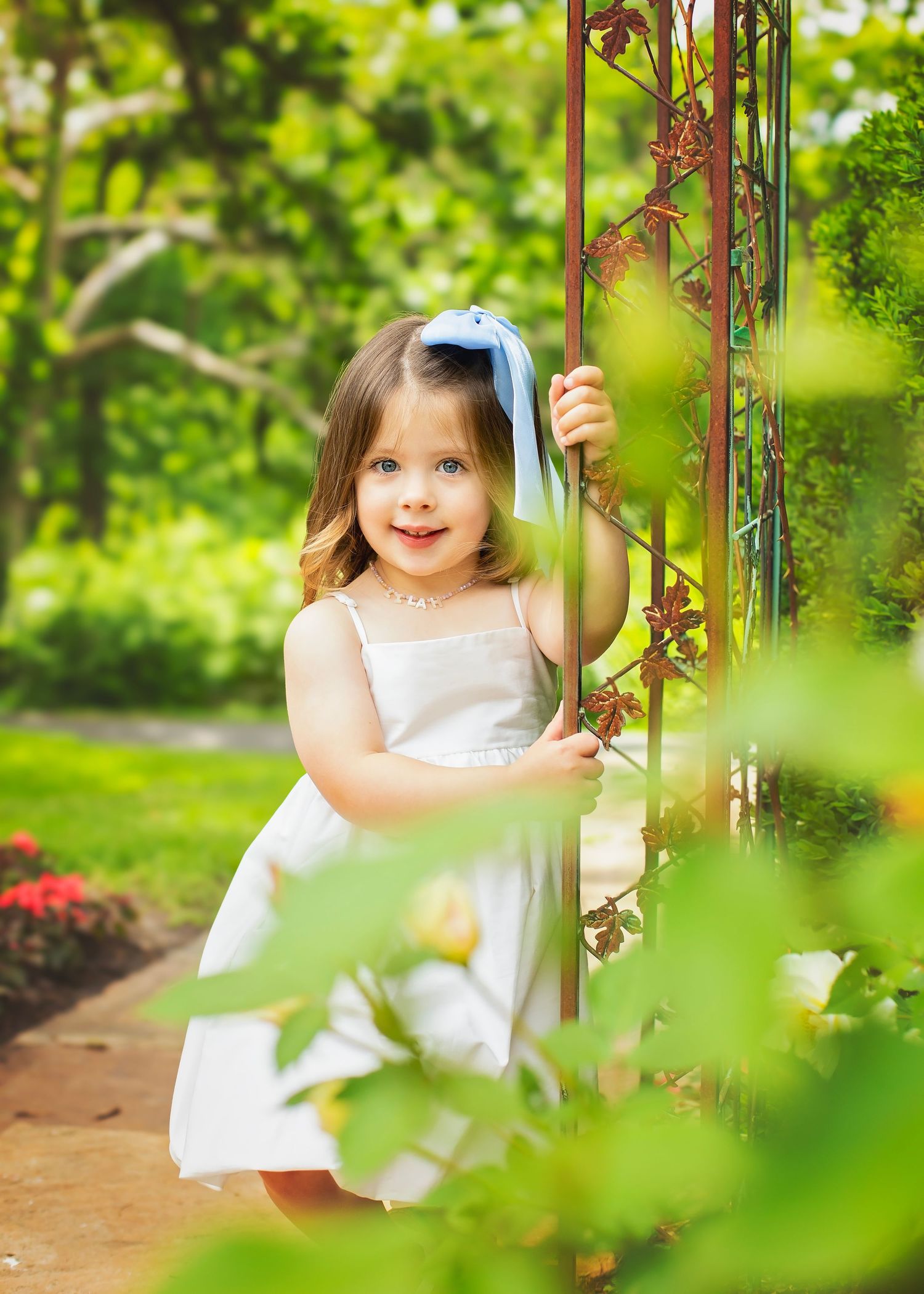 Charming little girl in white dress with blue bow smiling while holding garden trellis outdoors.