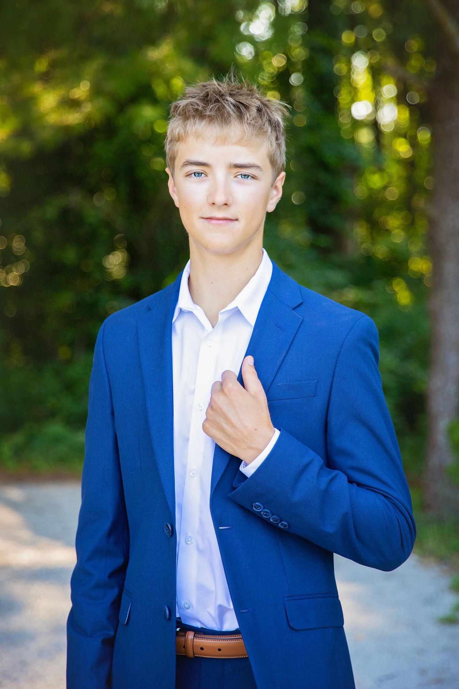 Handsome teenage boy in a blue suit jacket posing outdoors on a tree-lined path.