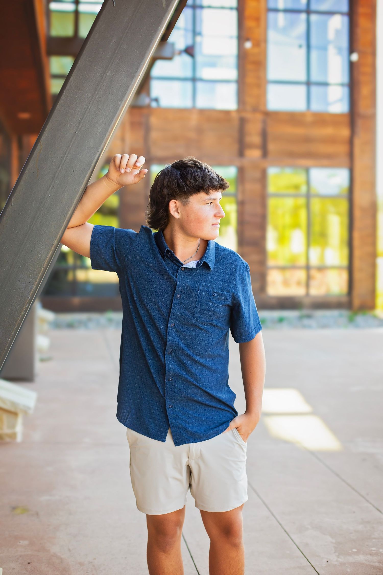 Teen boy in blue shirt leaning on a metal beam gazing sideways near a rustic wooden building.