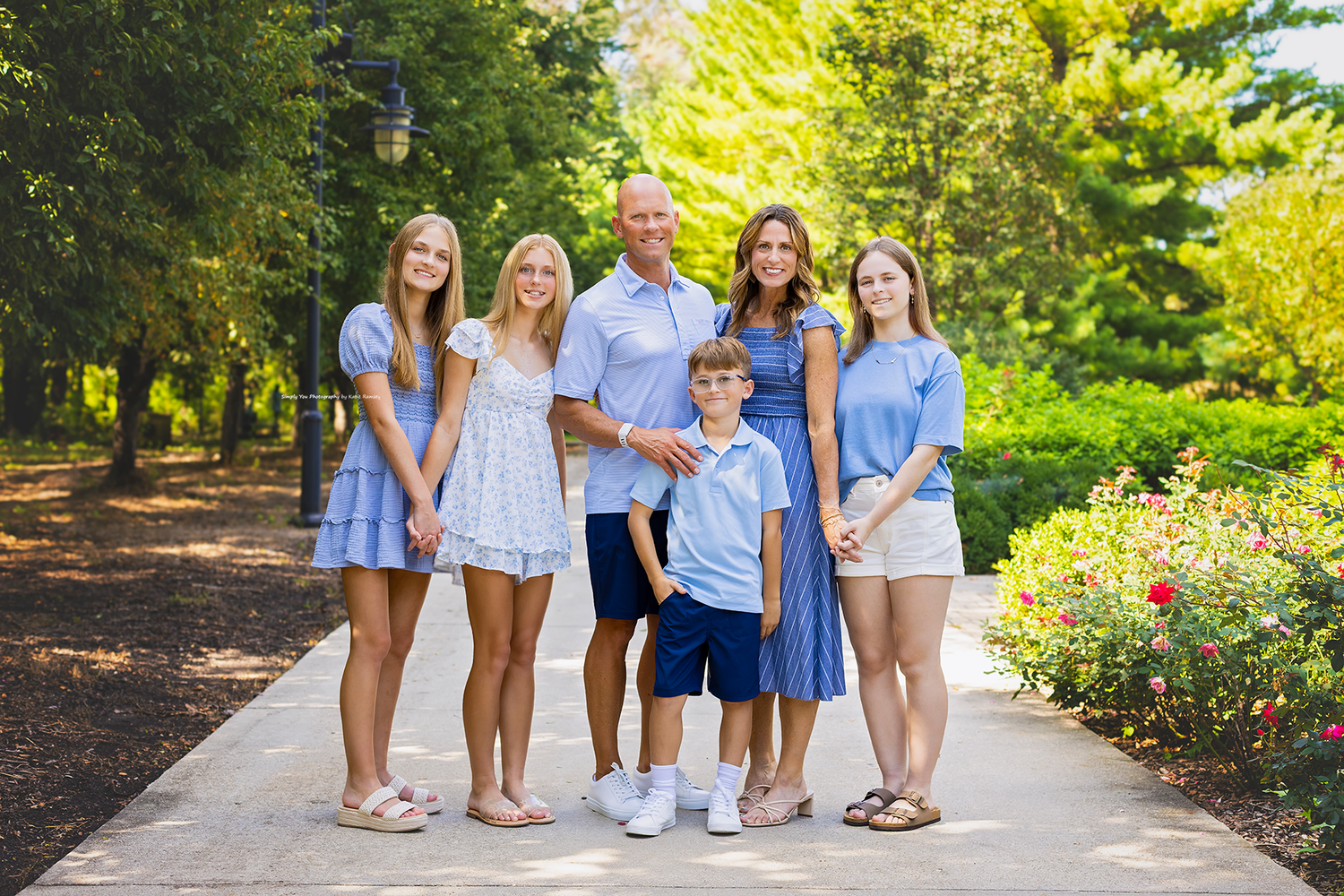 Family of six posing on a garden path, dressed in coordinating blue and white outfits, surrounded by lush green trees.