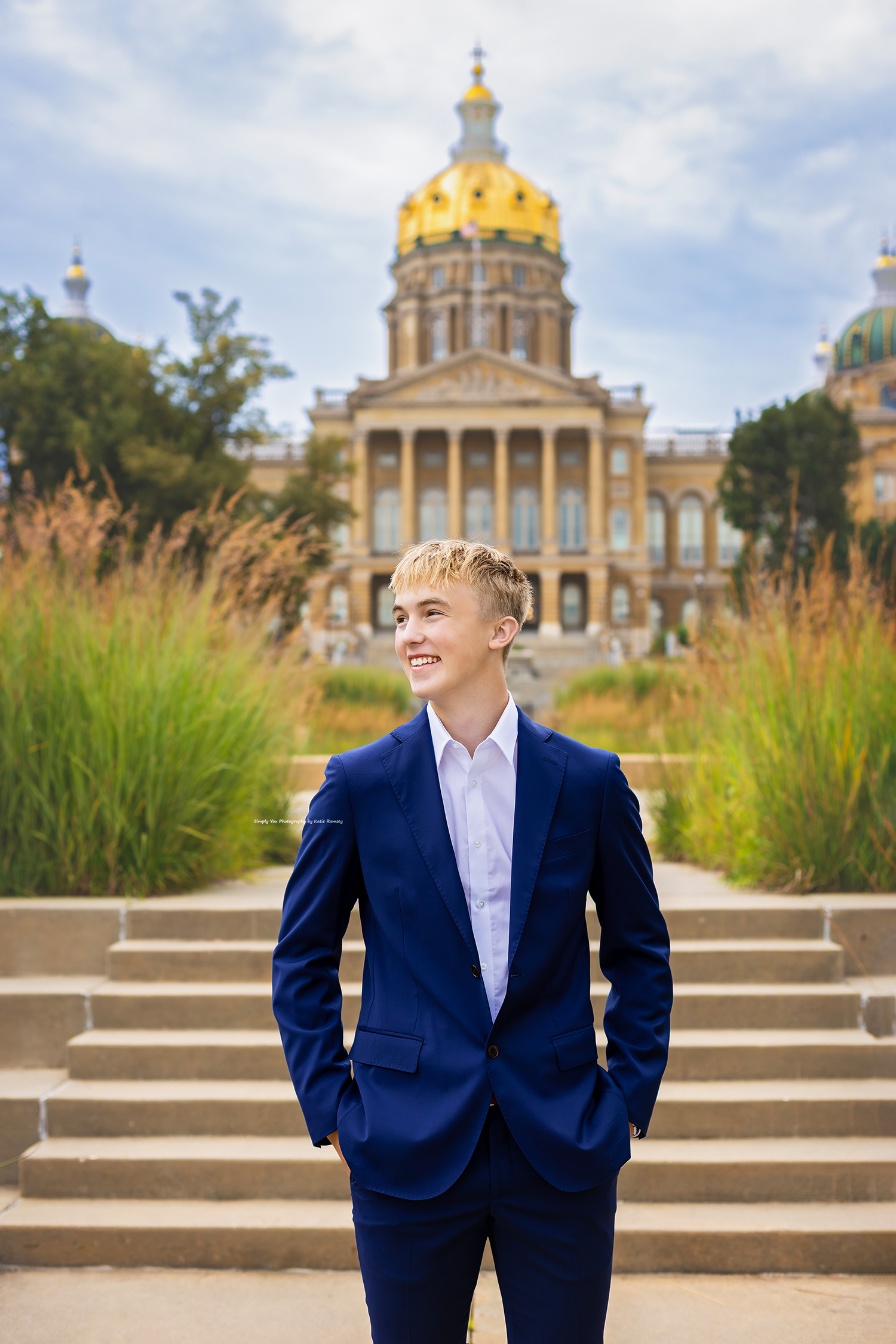 Young man in blue suit smiling in front of Iowa State Capitol building with golden dome.