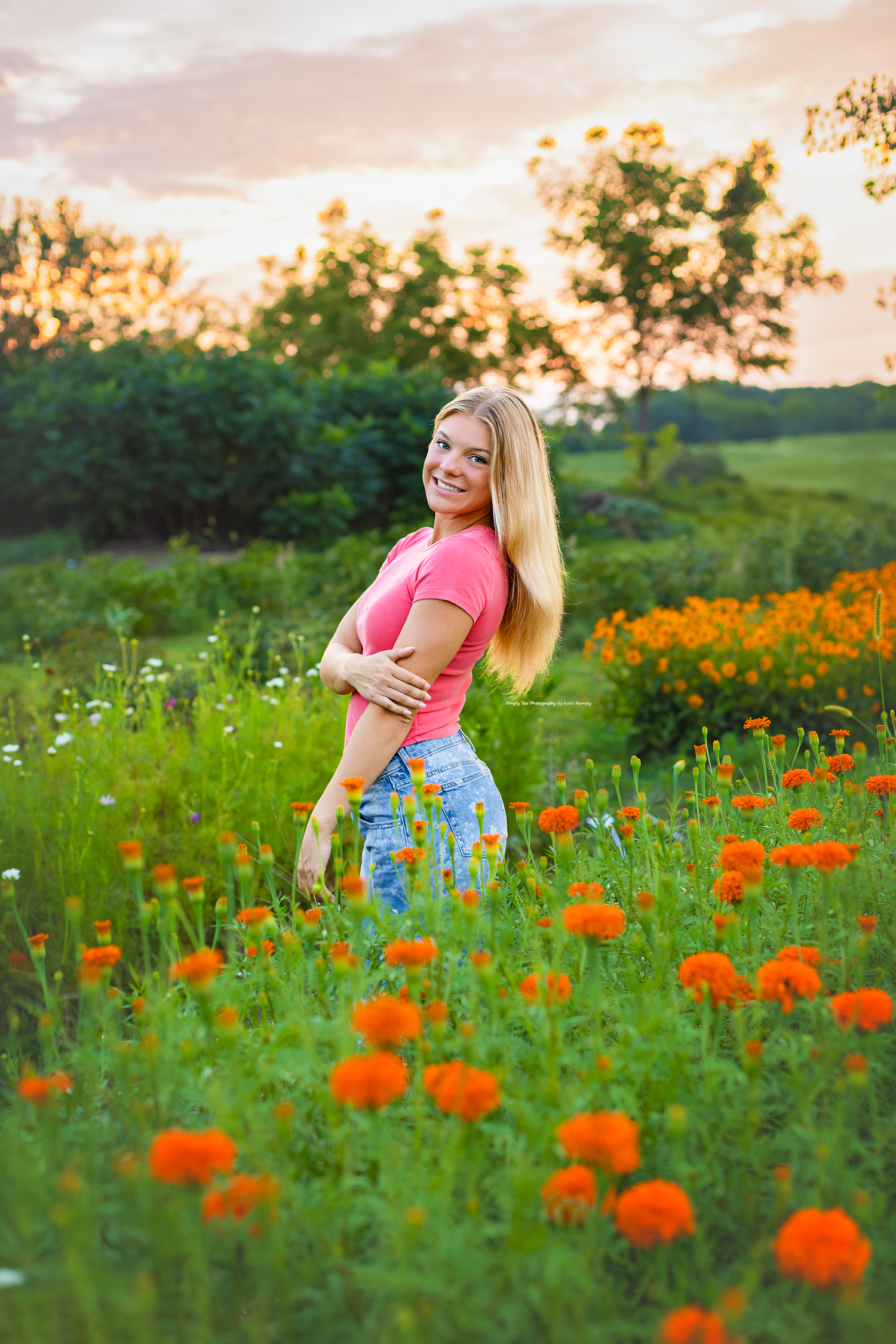Smiling blonde woman in pink top and floral jeans standing in a vibrant field of orange marigolds at sunset.