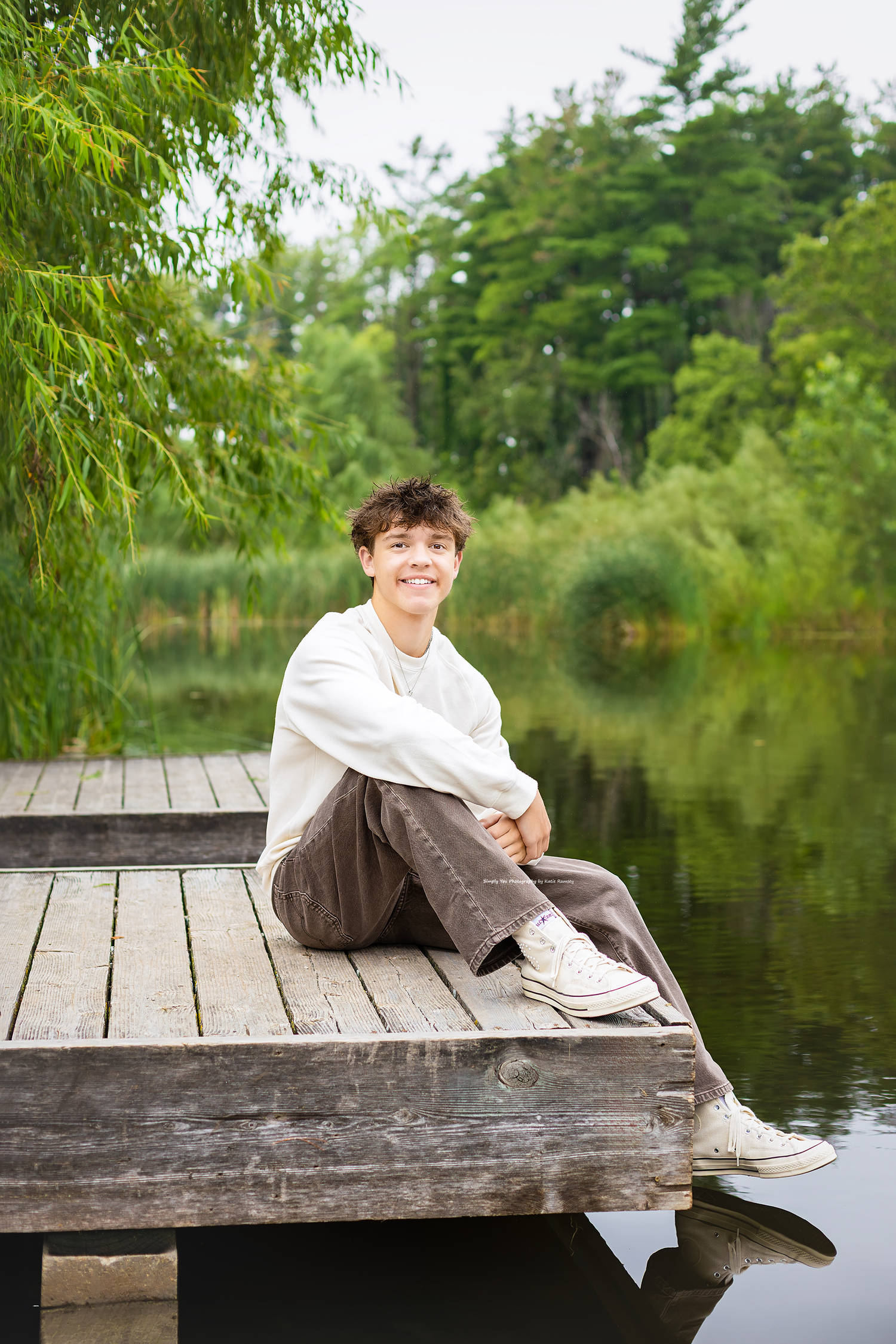 A smiling young man sits on a wooden dock by a serene pond, surrounded by lush green trees and willow branches.