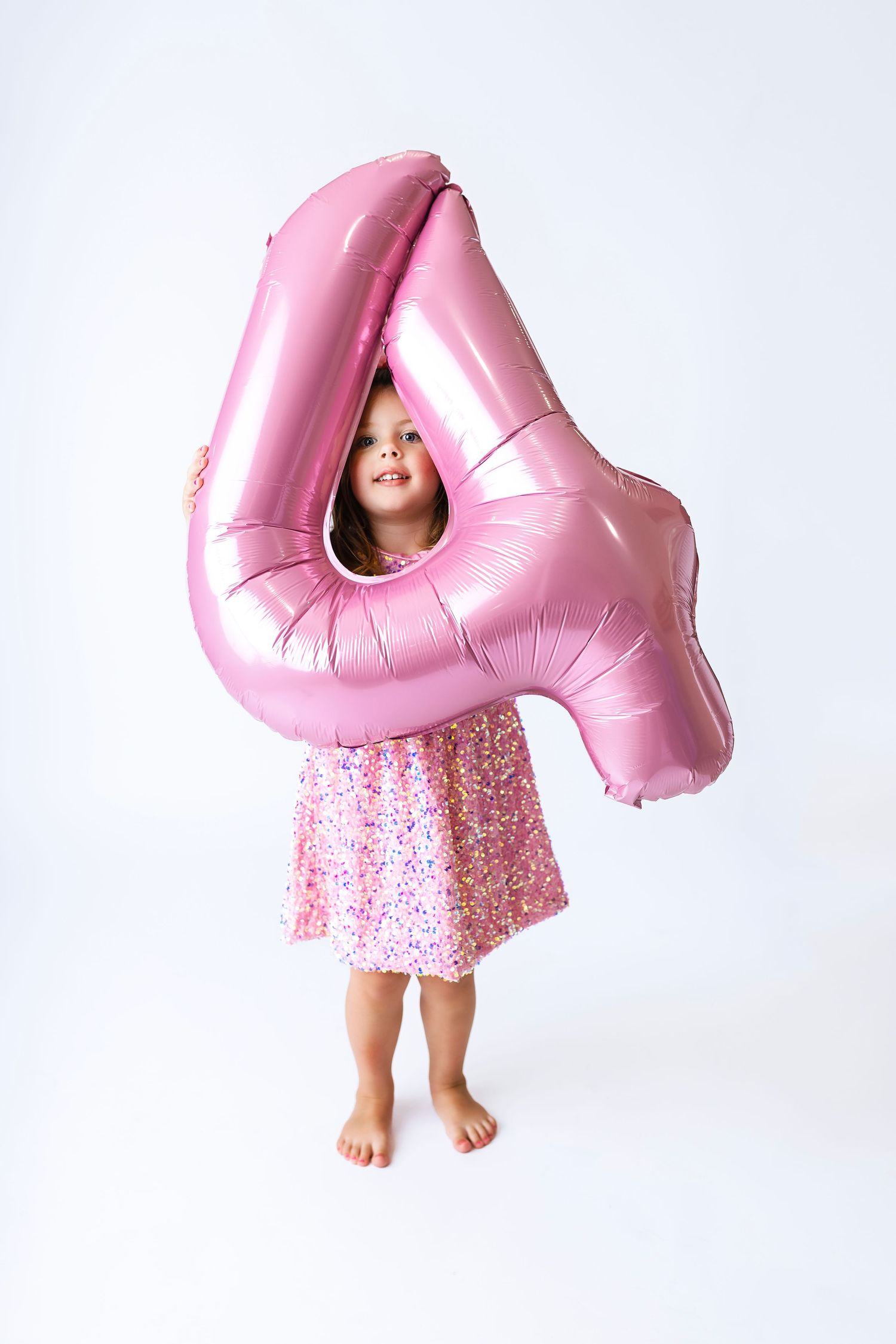 A young girl in a pink floral dress holds a large pink number 4 balloon on a white background.