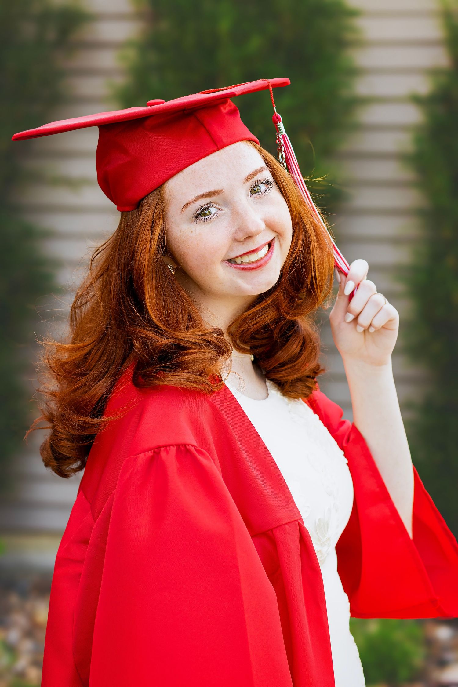 Happy redhead young woman in red graduation cap and gown smiling outdoors.