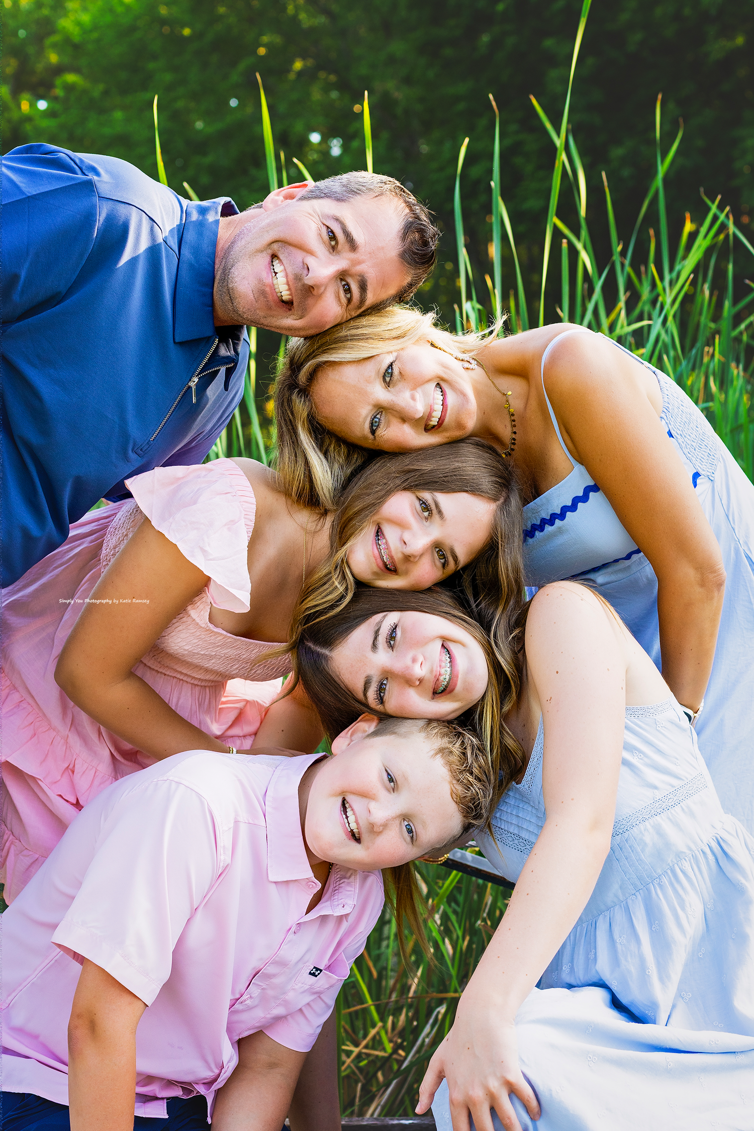 Happy family of five smiling together outdoors, leaning heads in, surrounded by lush green nature.