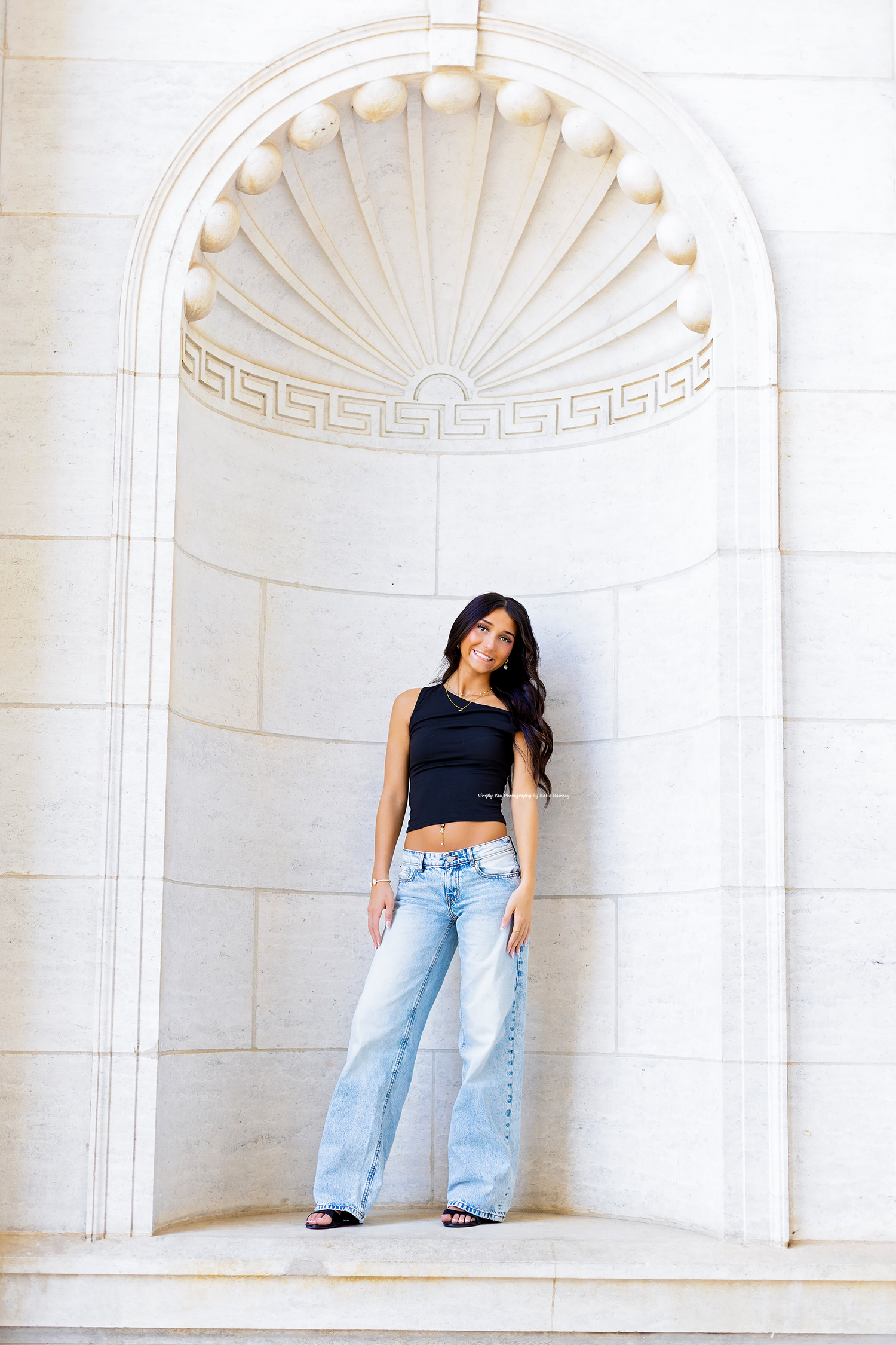Young woman in black crop top and wide-leg jeans posing in an ornate white stone archway.