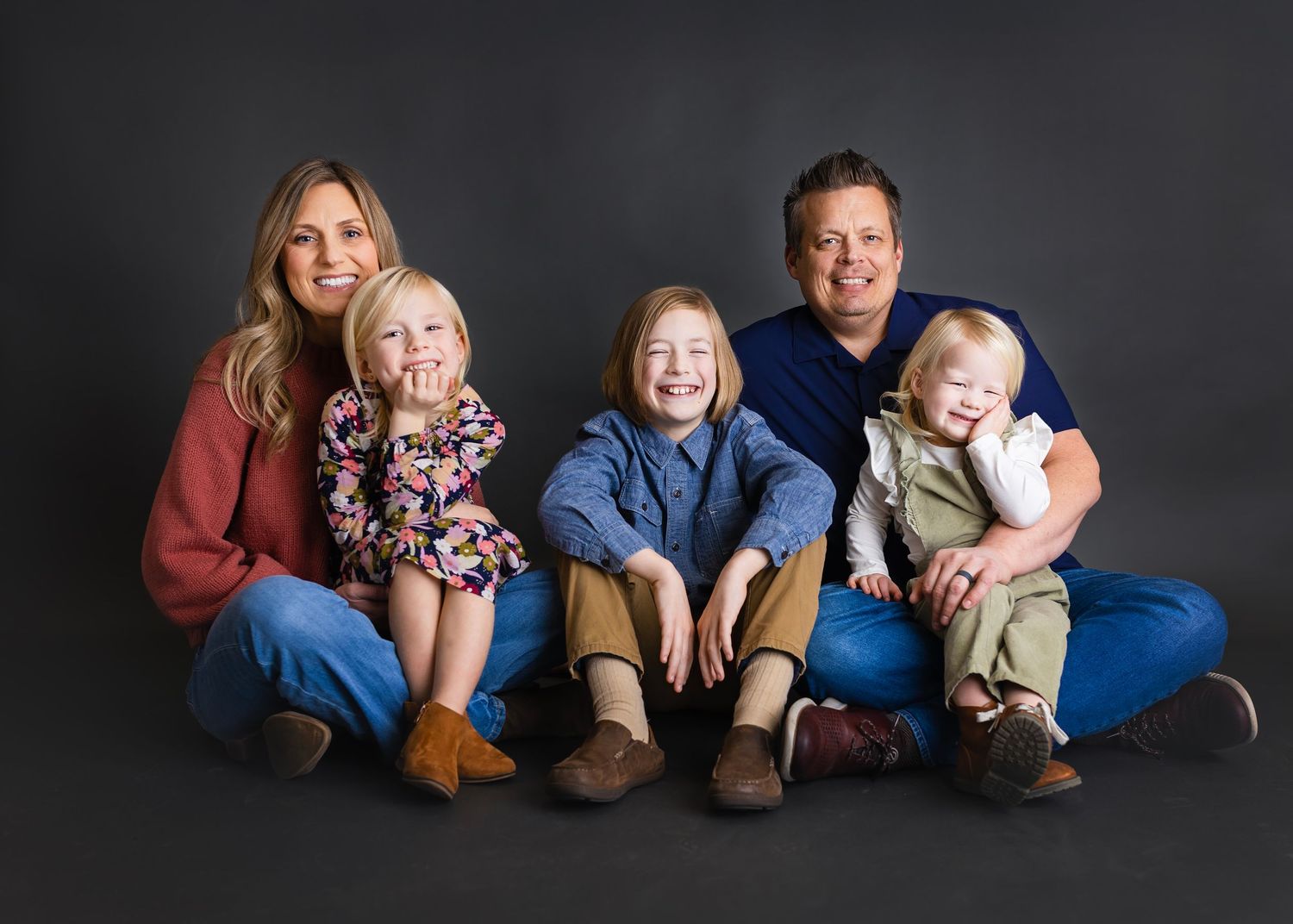 Happy family of five sitting together on floor, smiling, with two parents and three young children in studio portrait.