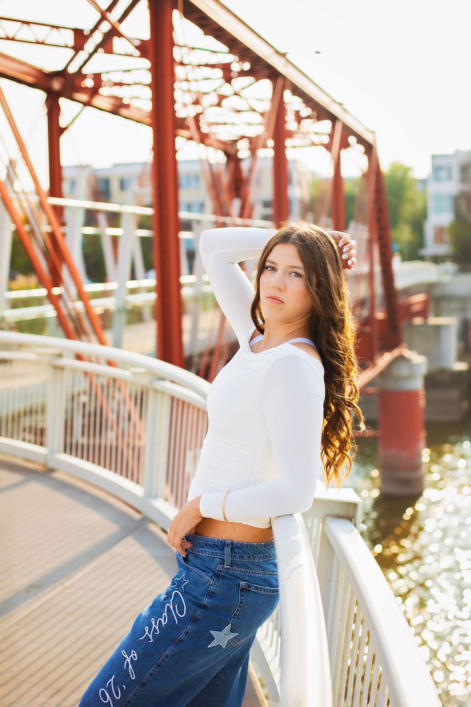Teen girl in white crop top and jeans poses on bridge over sparkling water.