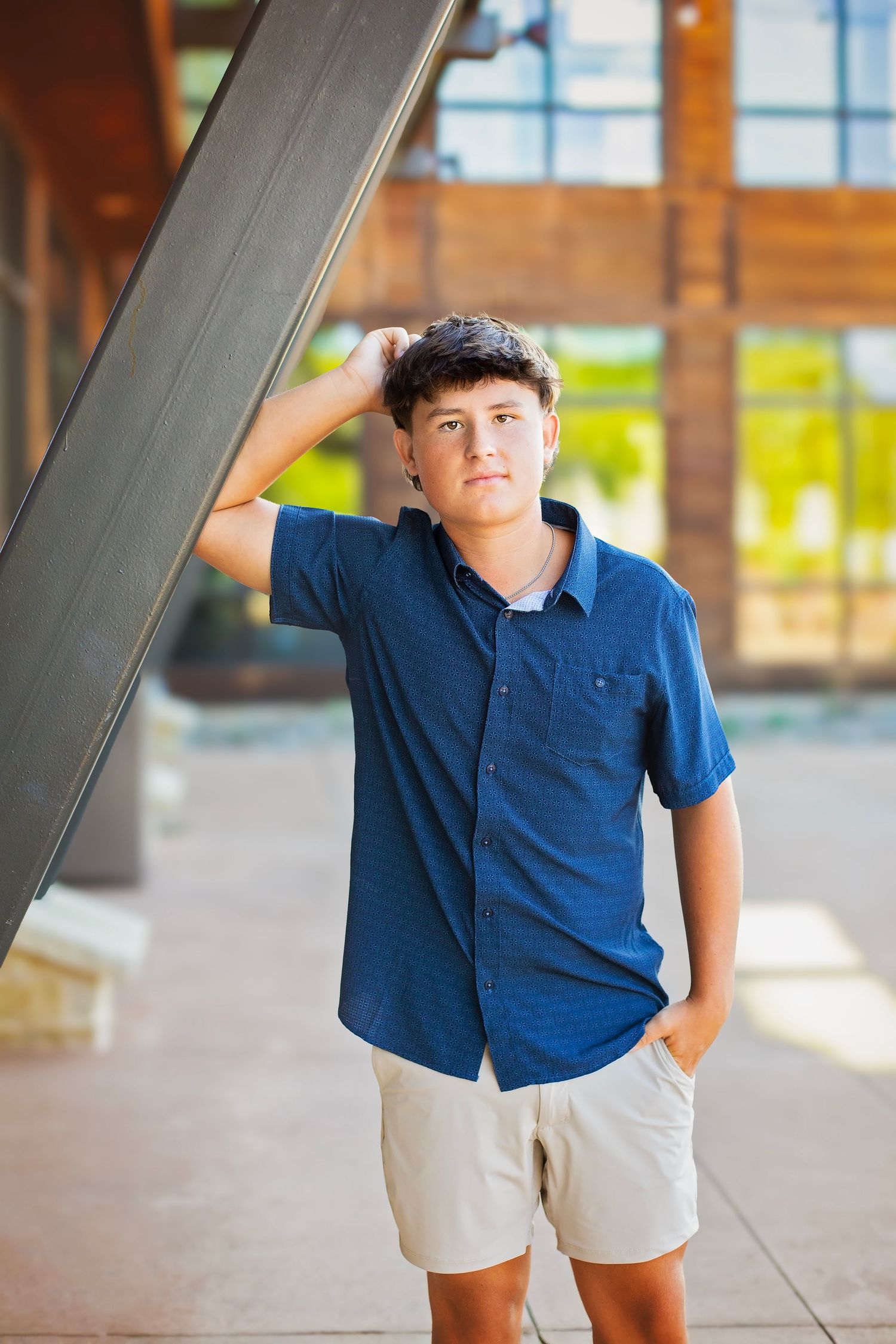 Teen boy in blue shirt and khaki shorts leaning against a metal beam outdoors during golden hour.