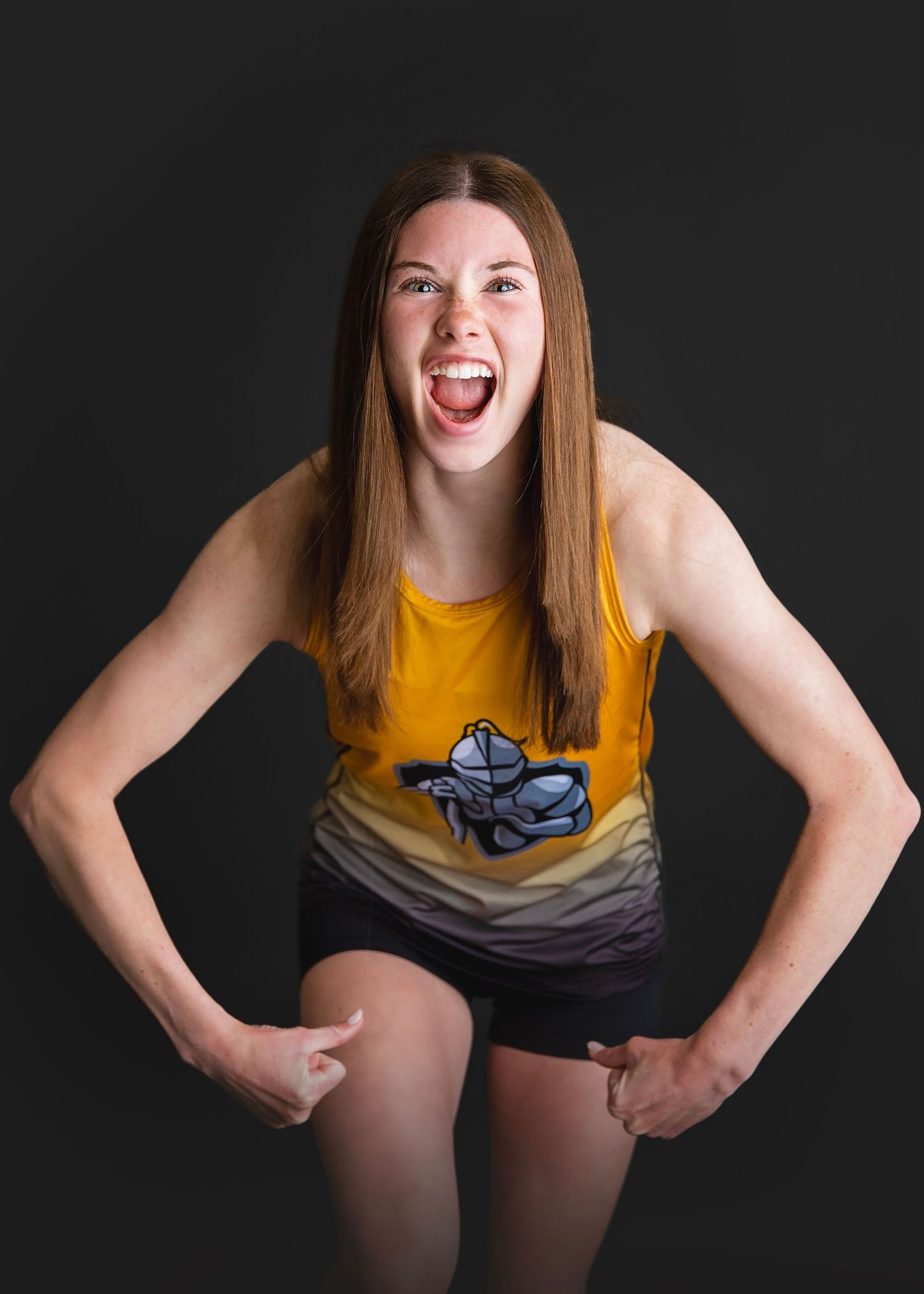 Excited teenage girl in yellow tank top with hands on knees laughing against dark background.