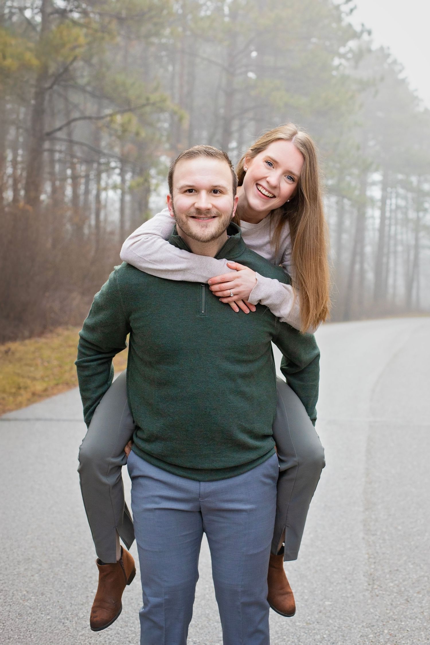 A smiling couple poses outdoors on a misty road, the man giving the woman a piggyback ride in a forest setting.