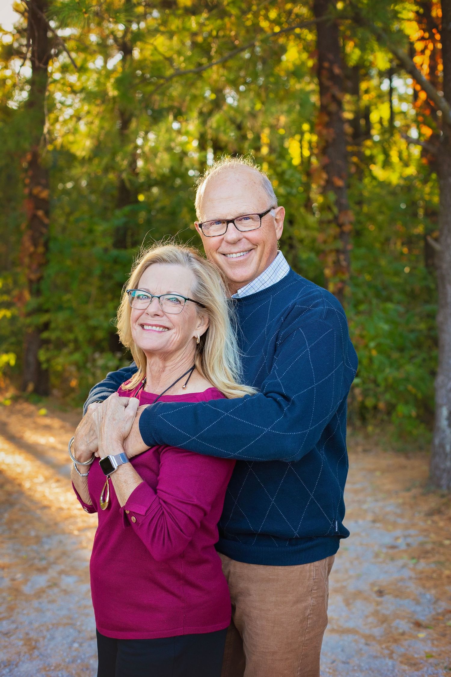Smiling older couple embracing warmly on a sunlit forest path in fall.