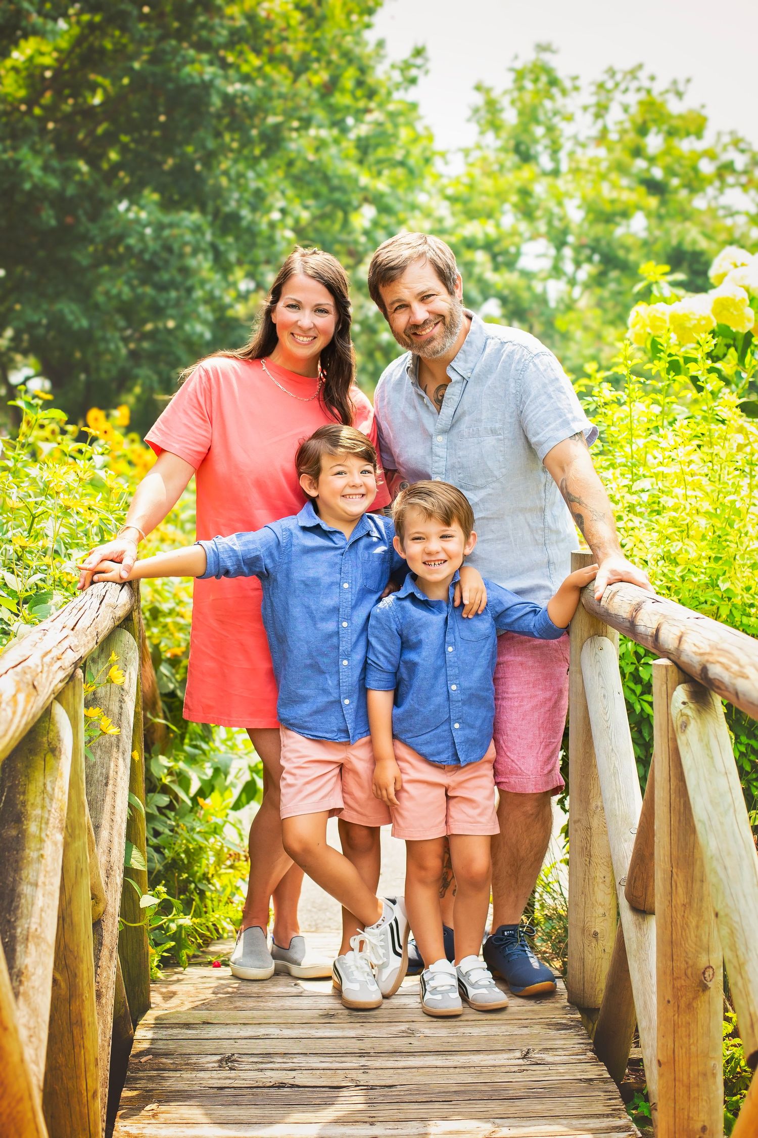 Smiling family of four poses together on wooden garden bridge surrounded by bright summer flowers.