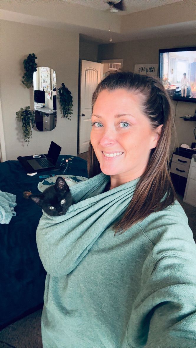 Smiling woman with blue eyes taking selfie with black cat perched on her shoulder indoors.