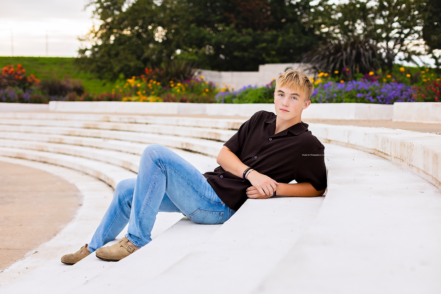 Teen boy in jeans and black shirt poses on white marble steps with colorful garden backdrop.