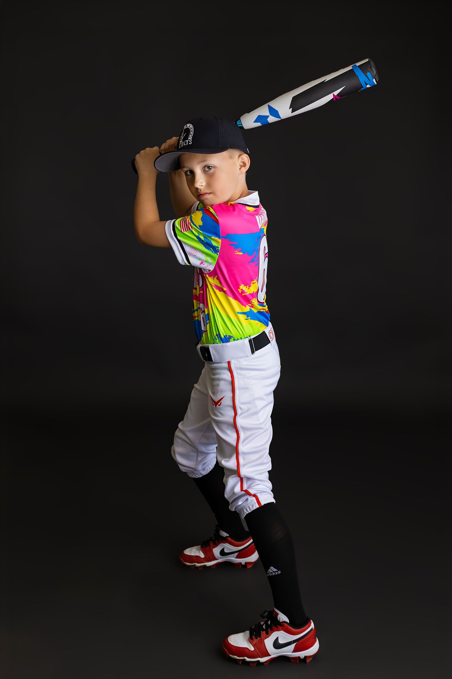 Young baseball player in colorful tie-dye jersey swings bat wearing white pants and red Nike cleats against dark background.
