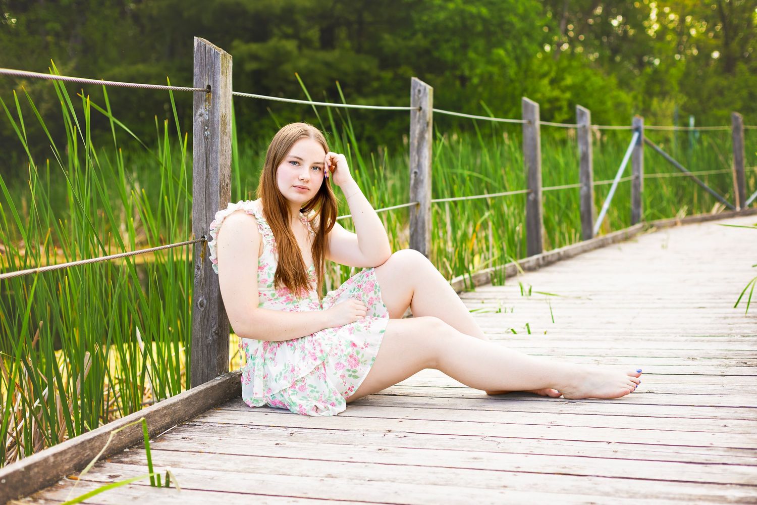 Young woman in floral dress sitting barefoot on wooden boardwalk surrounded by lush green reeds.