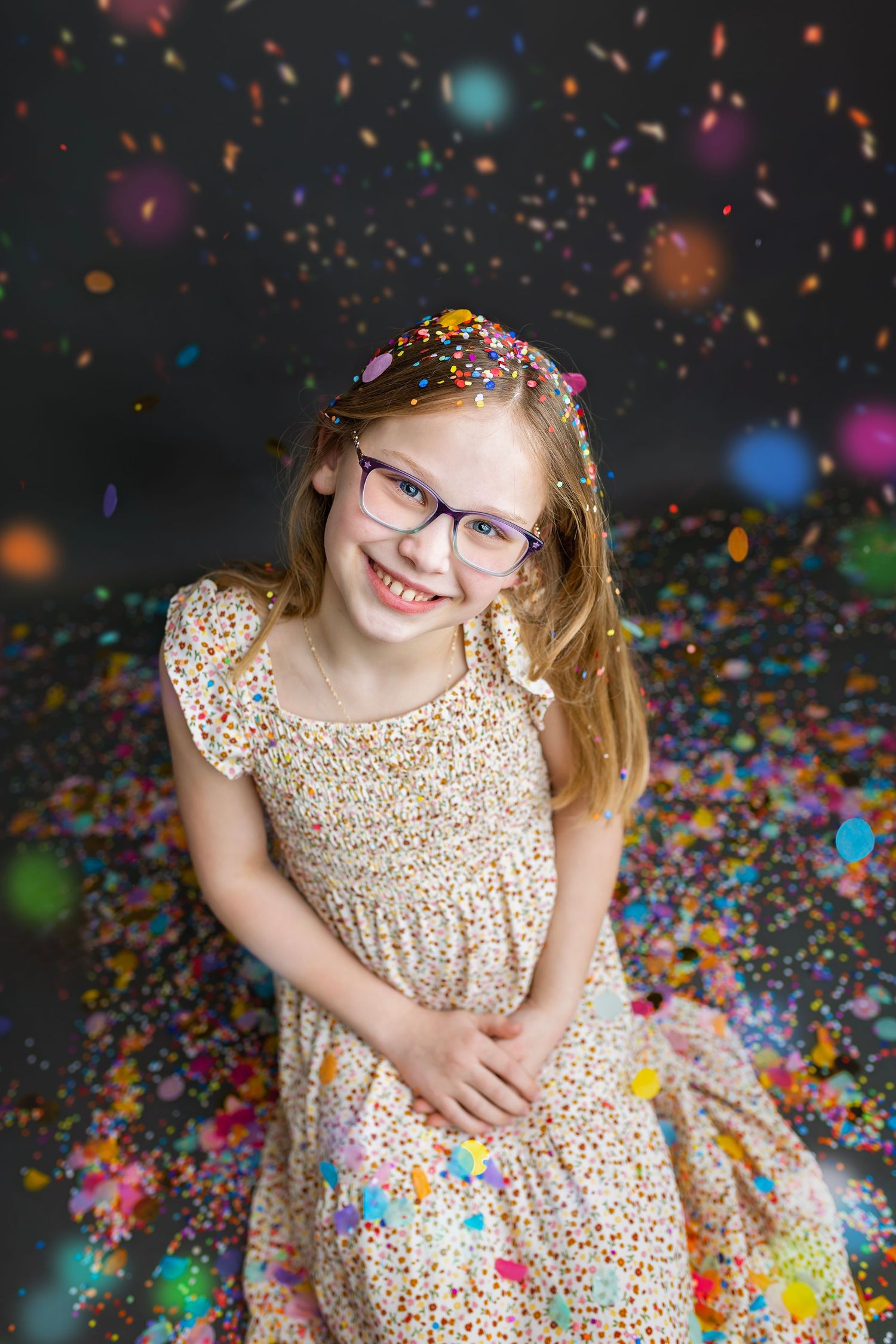 Cheerful girl in glasses and floral dress smiling among falling colorful confetti in studio.