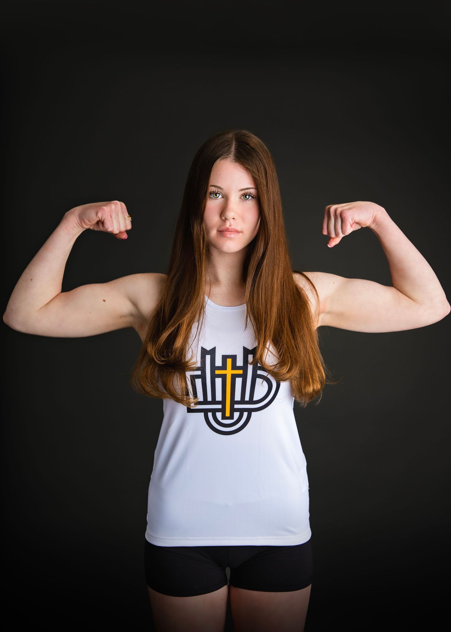 Young female athlete in white tank top flexes both biceps against a dark studio background.