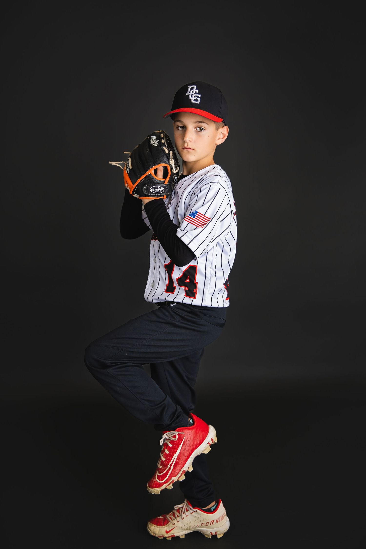 Young baseball pitcher in white pinstripe jersey number 14 with American flag patch poses in pitching stance with glove.
