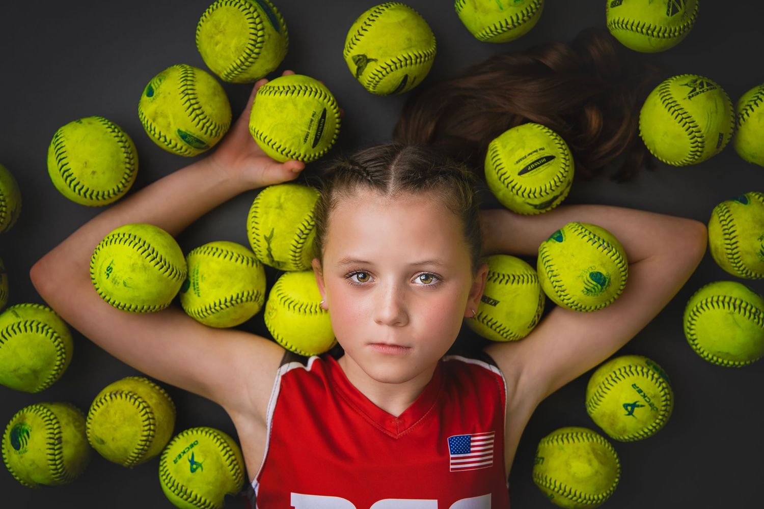 Young softball player lies surrounded by yellow softballs in creative studio sports portrait.