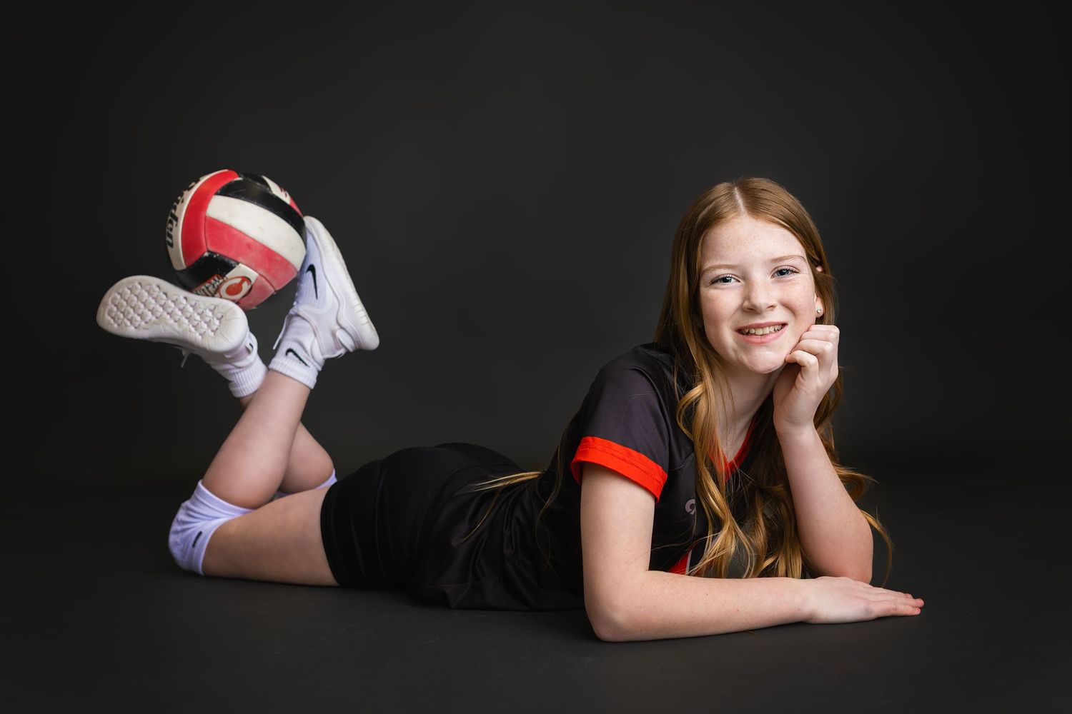 Young girl in black volleyball uniform lying on floor smiling with volleyball balanced on her feet.