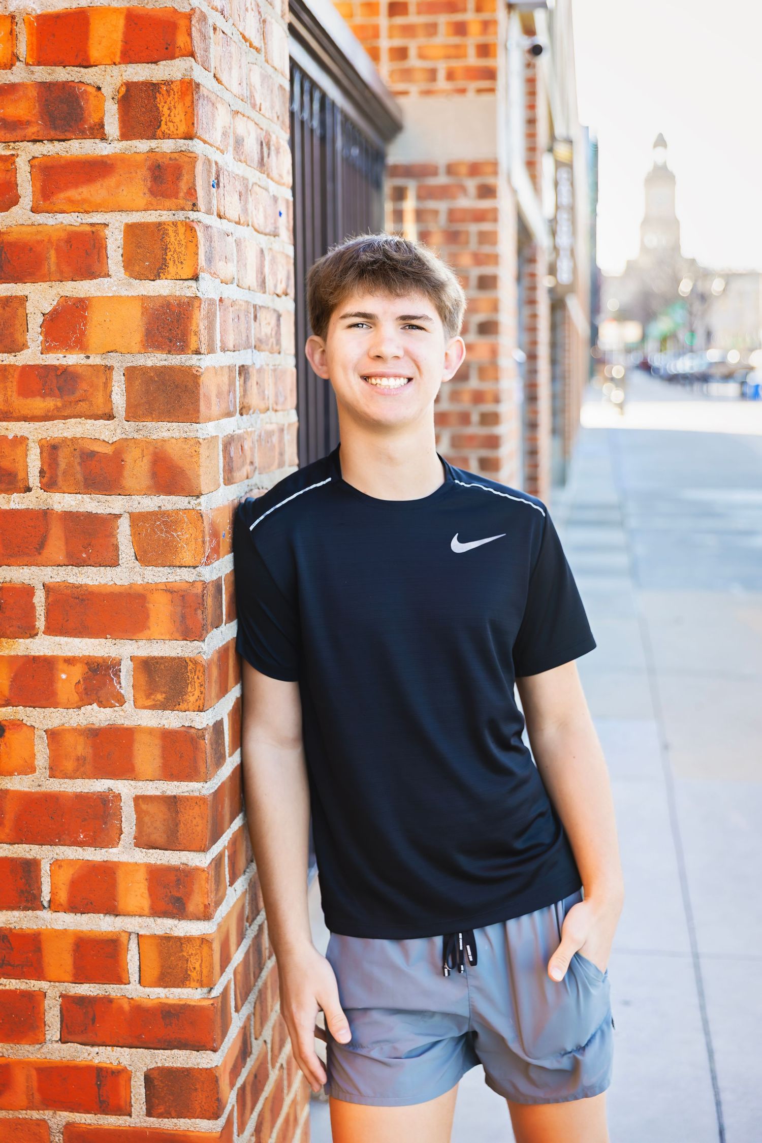 Teenage boy in black Nike shirt smiling while leaning against a red brick wall.
