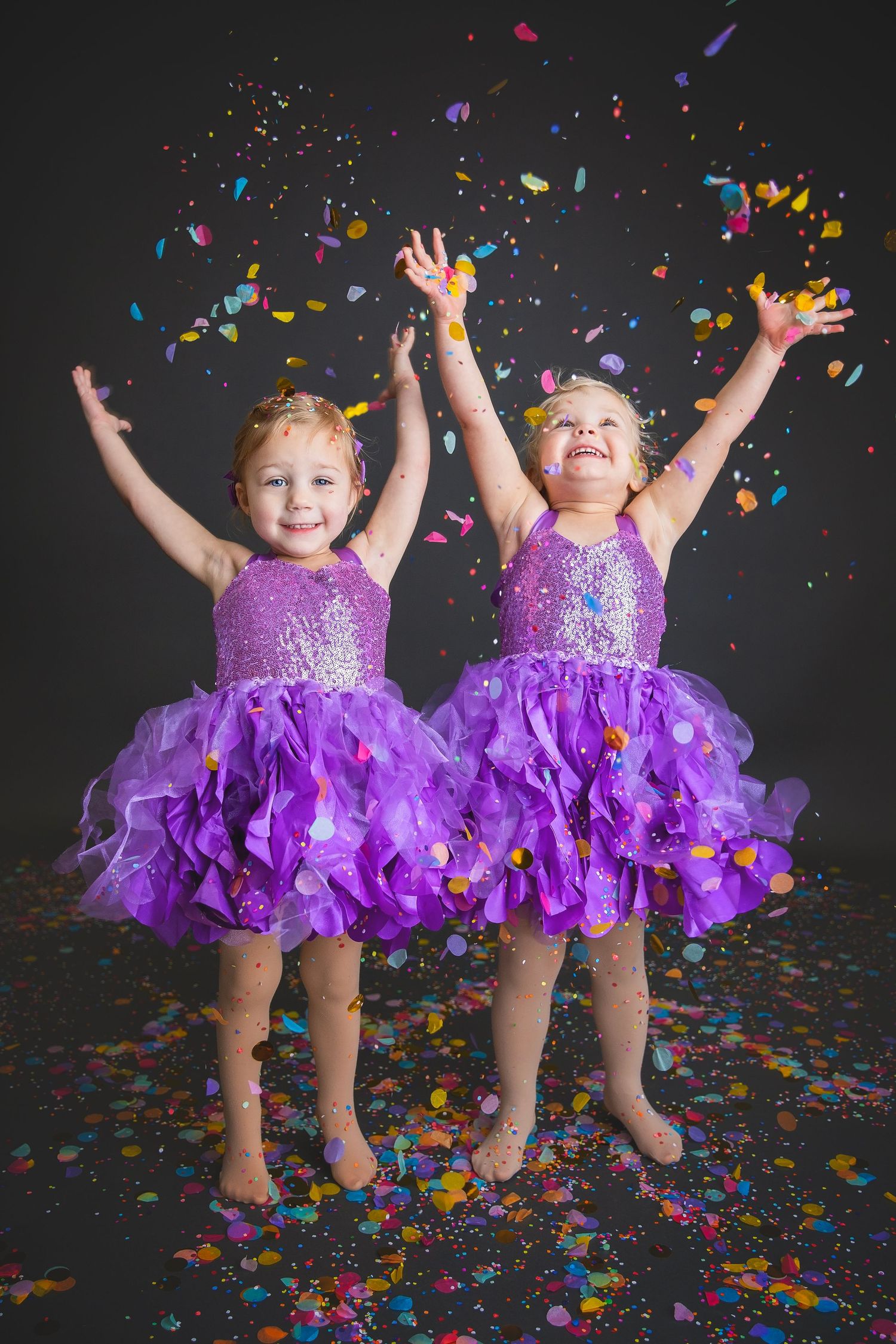 Two girls in matching purple sequin dresses jumping with colorful confetti on dark background.