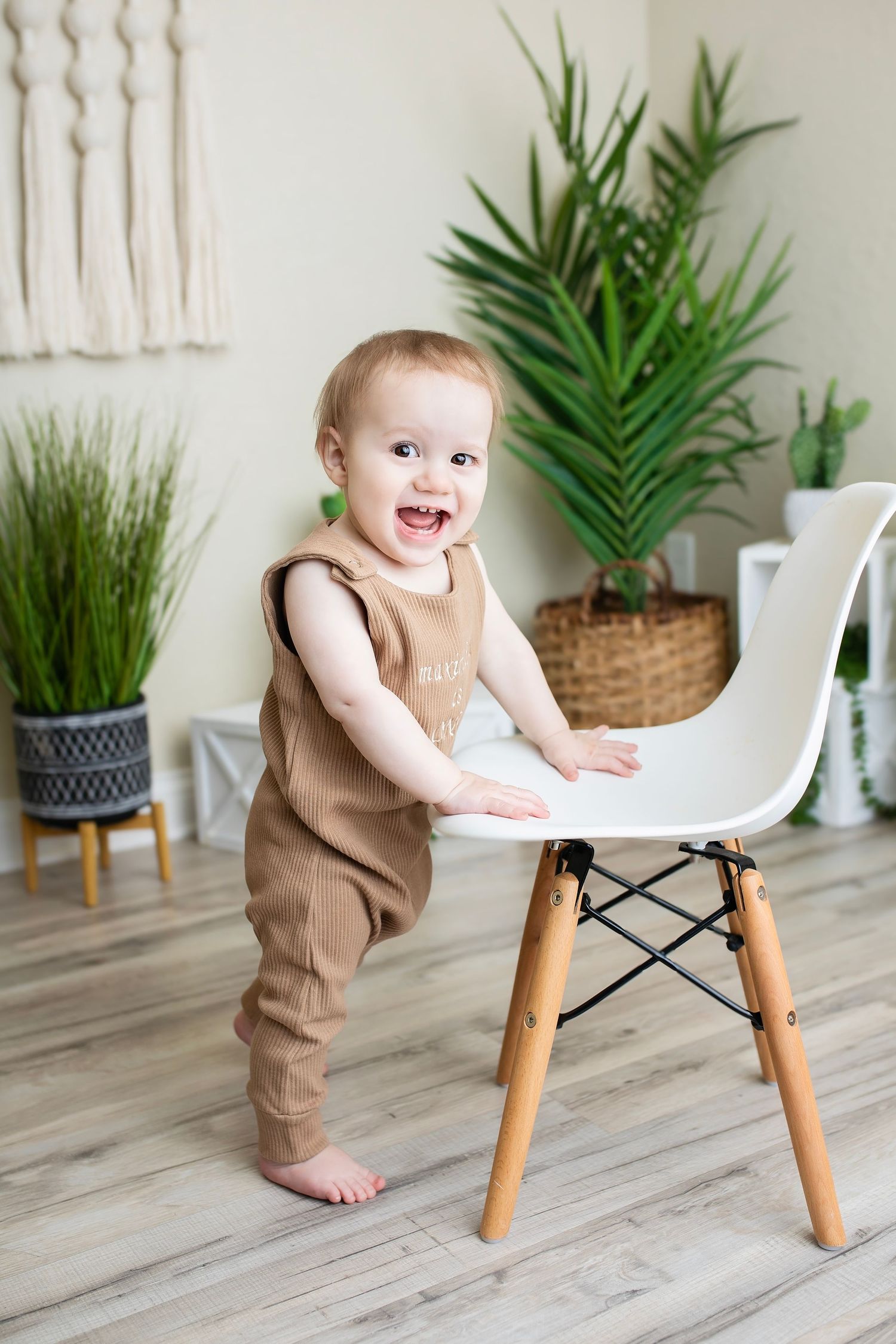 Happy baby boy in brown romper standing at a white modern chair with indoor plants in background.