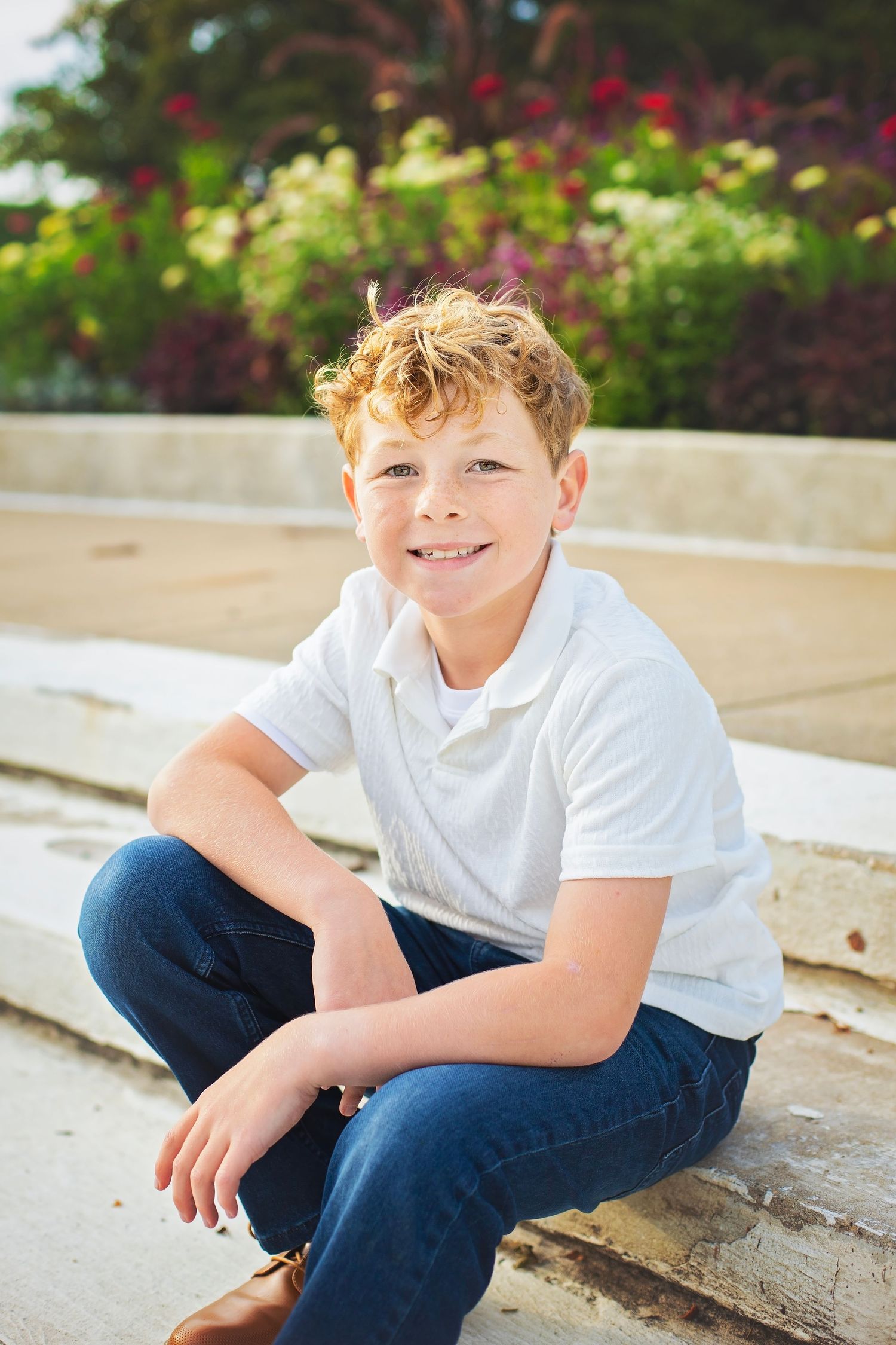 Happy red-haired boy smiling while sitting cross-legged on stone steps with flowers behind him.