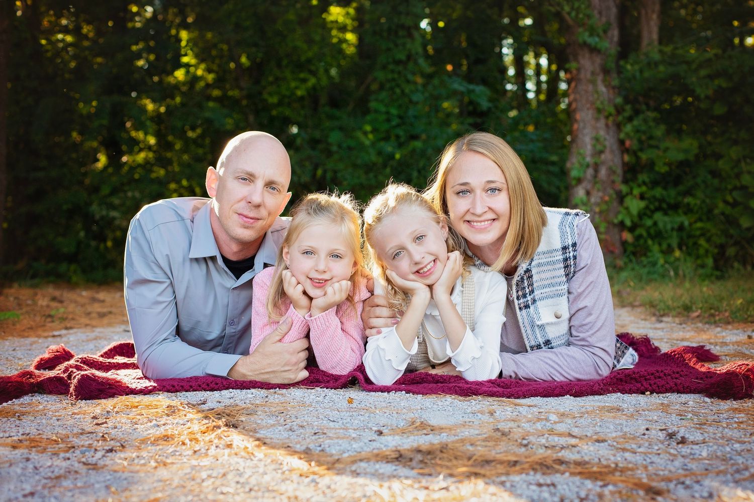 Family of four lying on a red blanket smiling at camera with trees in background.