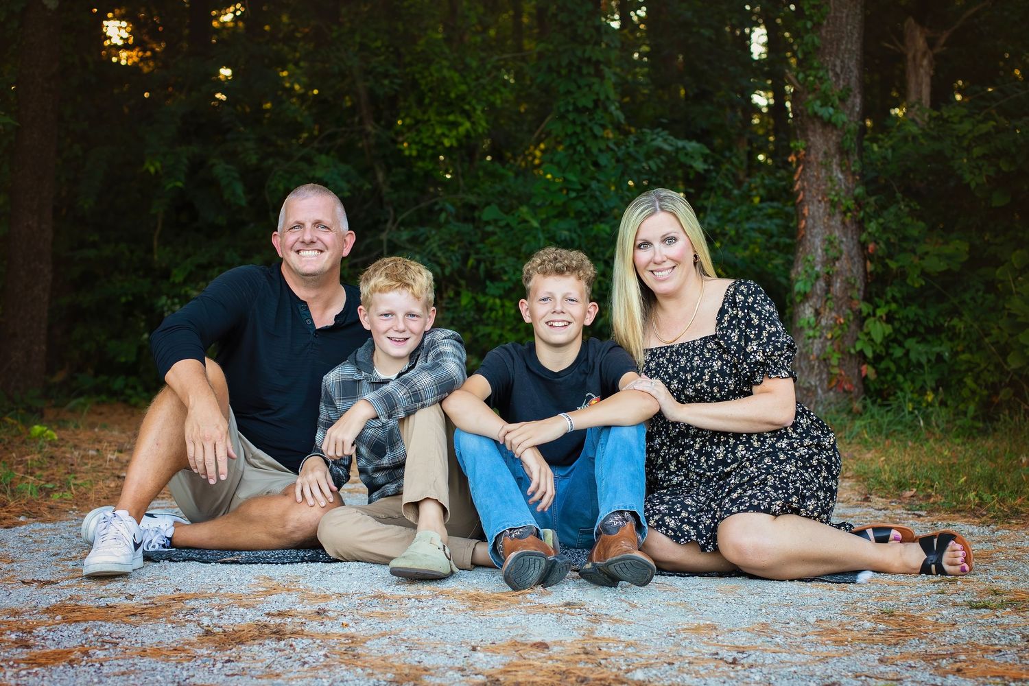 A cheerful family of four sits casually on the ground smiling together in front of a wooded backdrop.