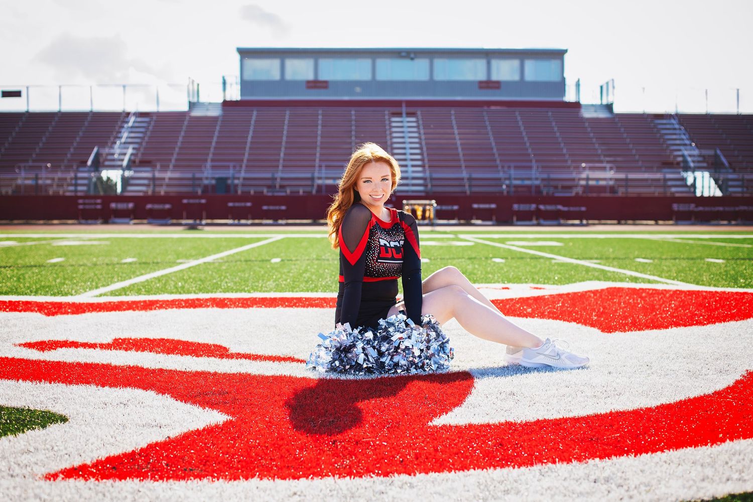 Young cheerleader in uniform poses with pom-poms on red and white logo at football stadium.